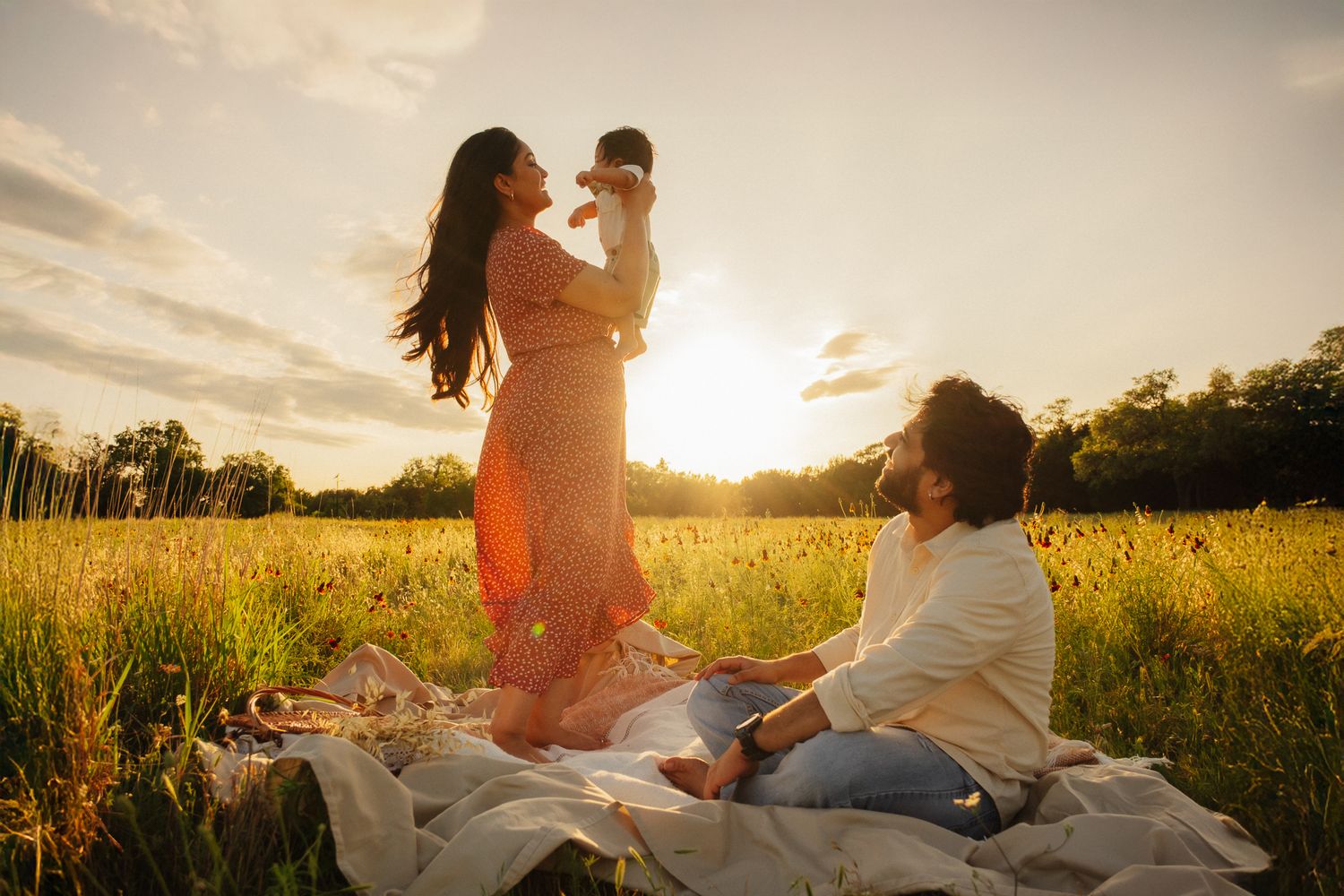 People sit on blanket in sunny meadow during golden hour with sunflare illuminating grass field at sunset.