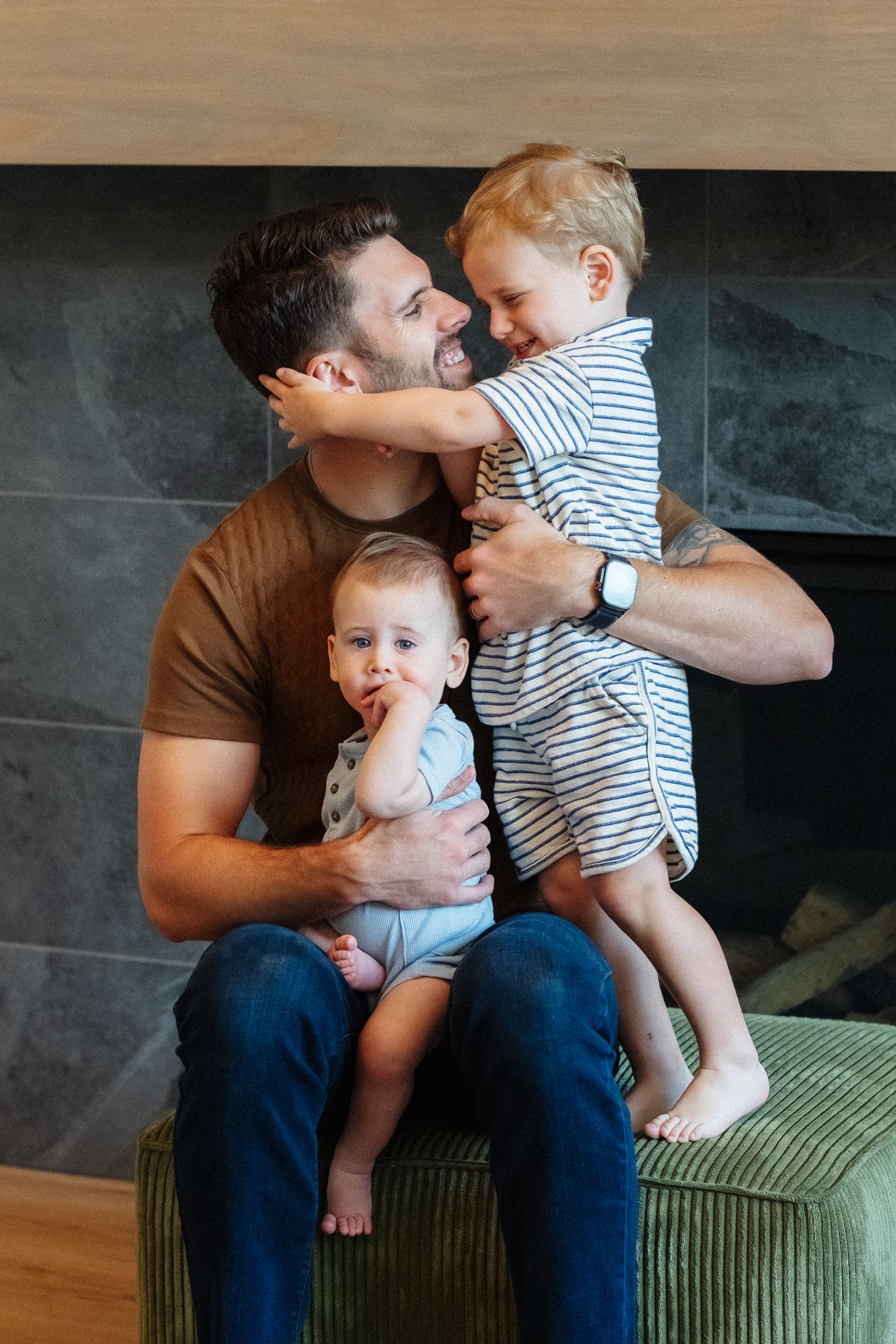 A tattooed father sits by a fireplace smiling as his toddler hugs his face while holding his baby on his lap.
