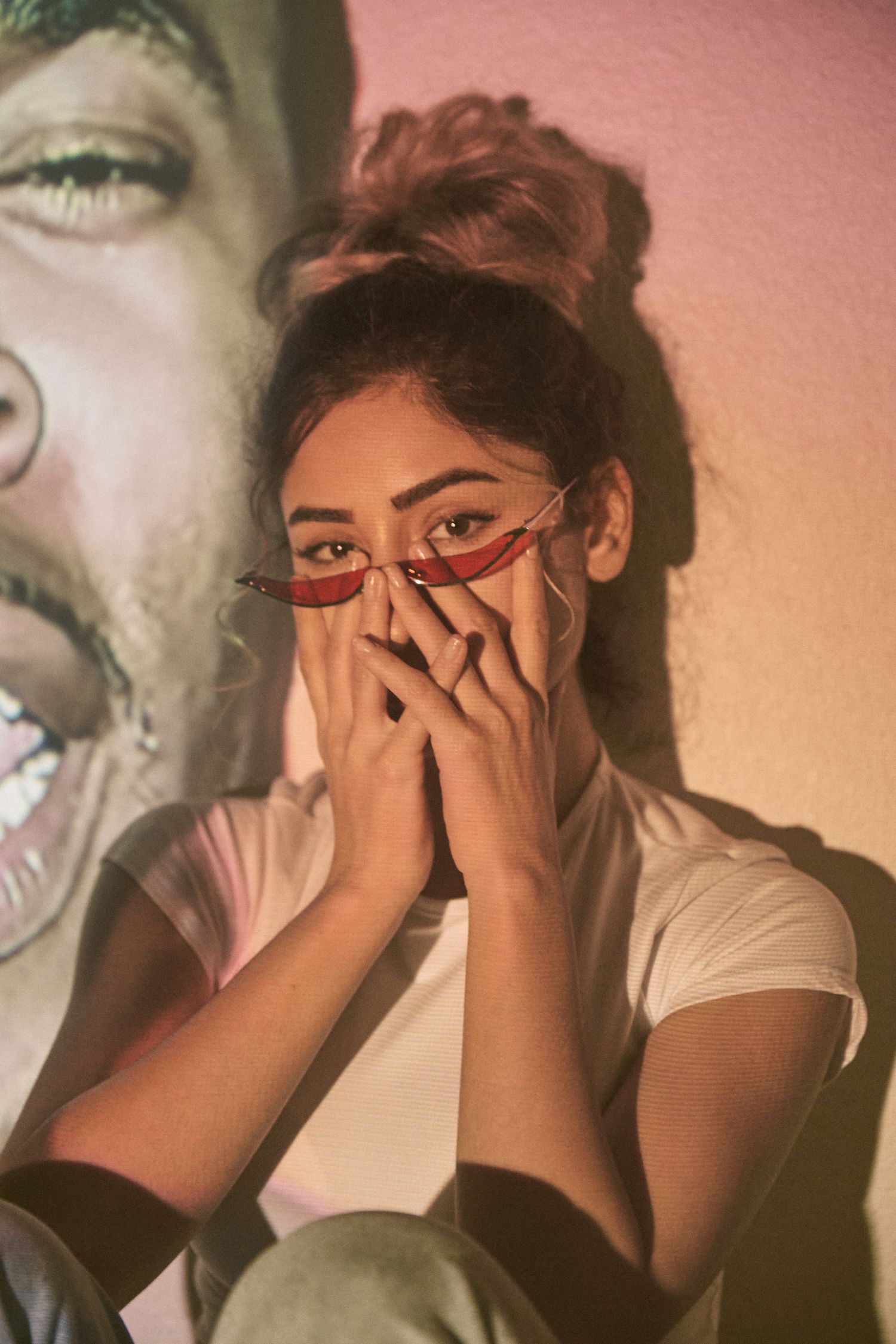 A person in red sunglasses and white t-shirt covers their mouth playfully while posing against a peachy backdrop.