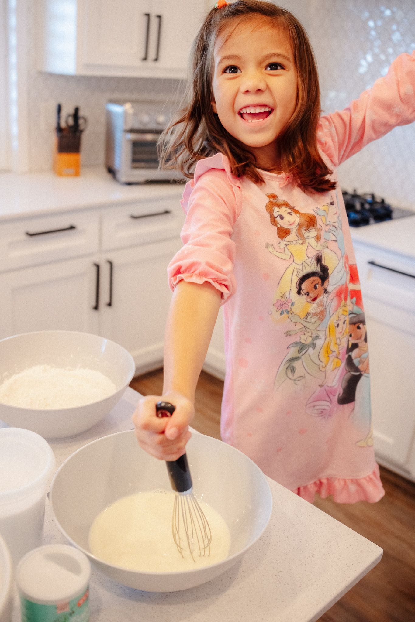 A child in a pink shirt enthusiastically mixing ingredients in a large white bowl on a kitchen counter.