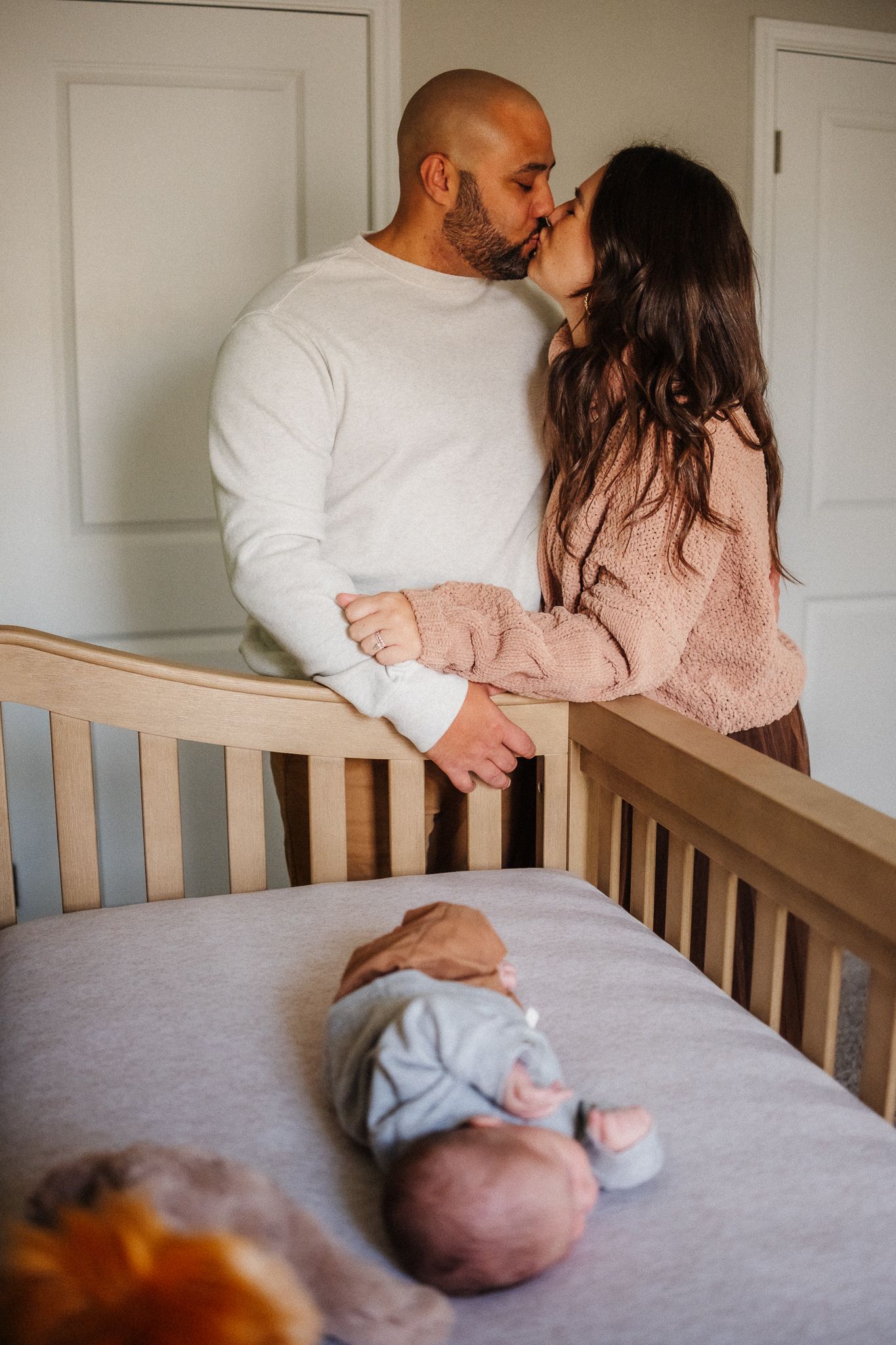Parents sharing a loving moment near a wooden crib with their newborn baby.