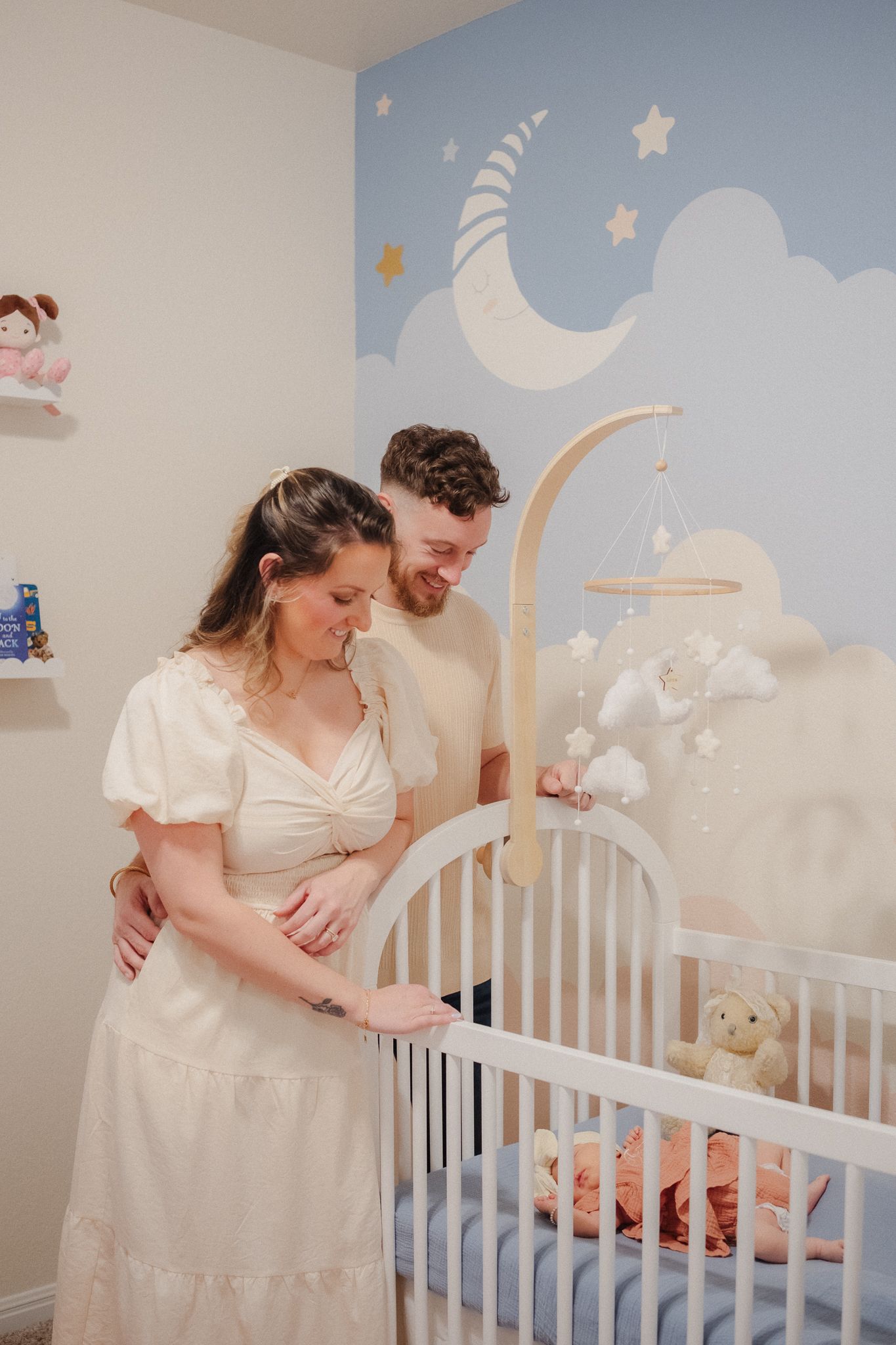 Expectant parents smile over a white crib in a soft nursery with a moon and stars mural.