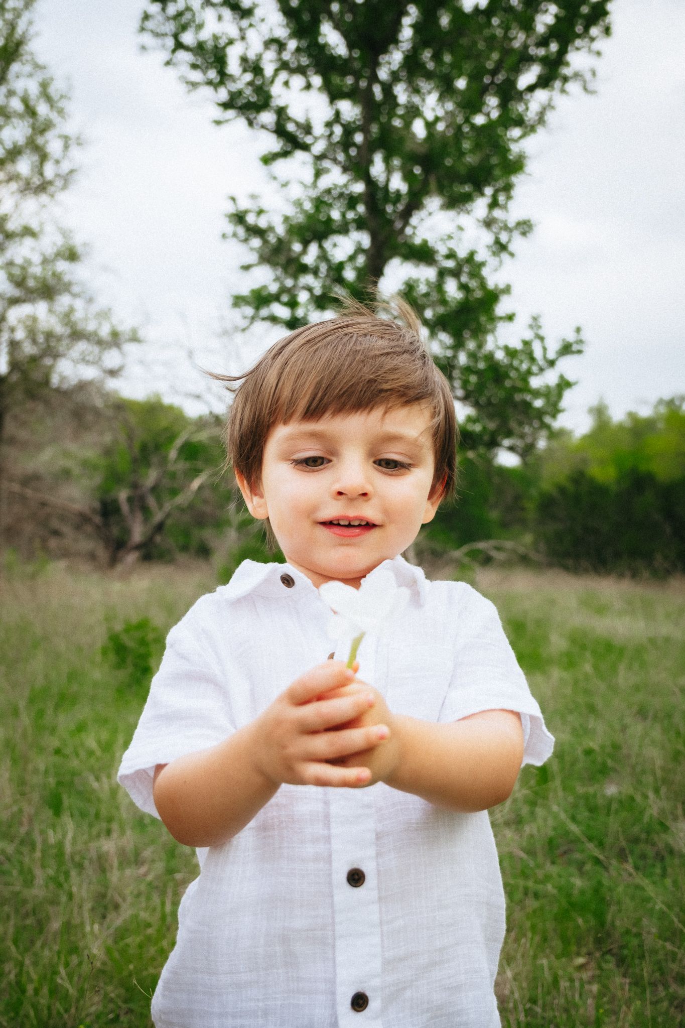 Young child wearing a white shirt laughs playfully while standing in a grassy meadow on a sunny day.
