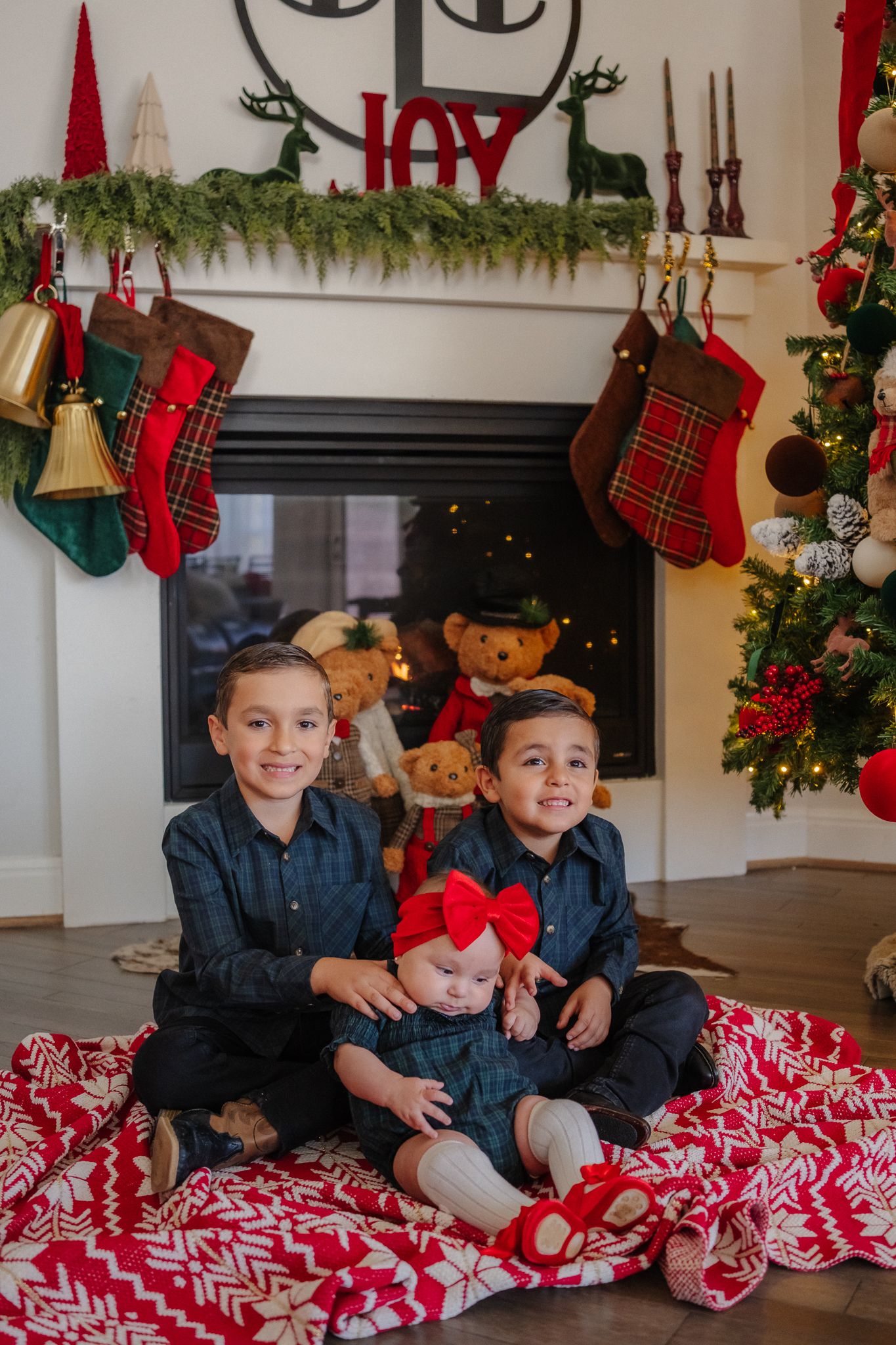 Three children in matching navy outfits pose in front of a decorated Christmas fireplace with stockings and garland.