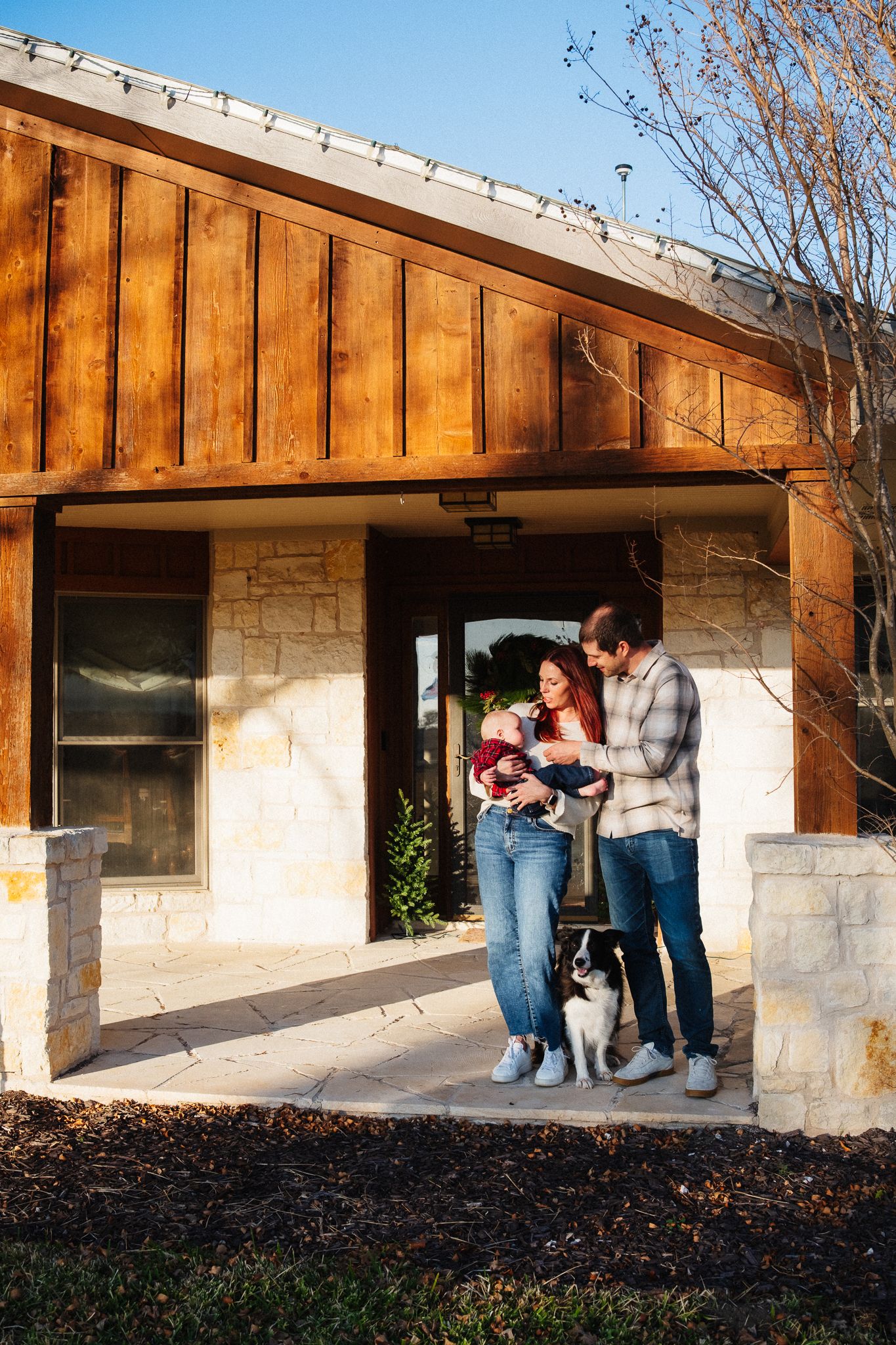 A happy family with a baby and dog stands on the porch of a modern stone and wood home.