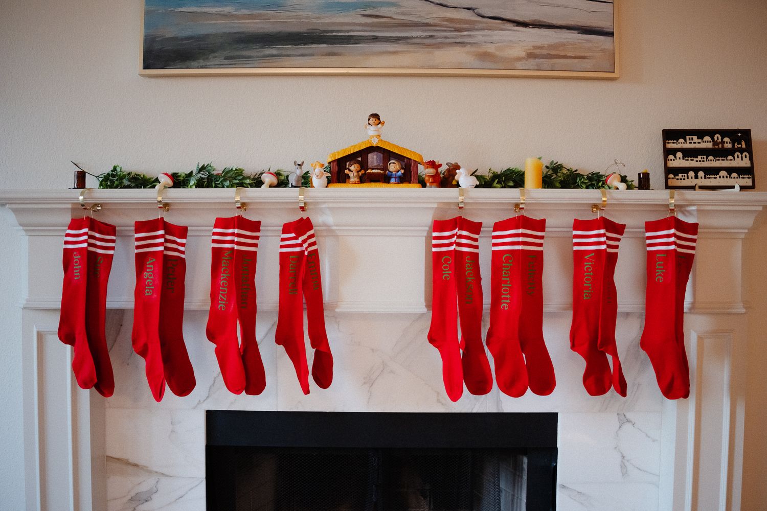 Nine red Christmas stockings hung on a white fireplace mantel with garland and a nativity scene.
