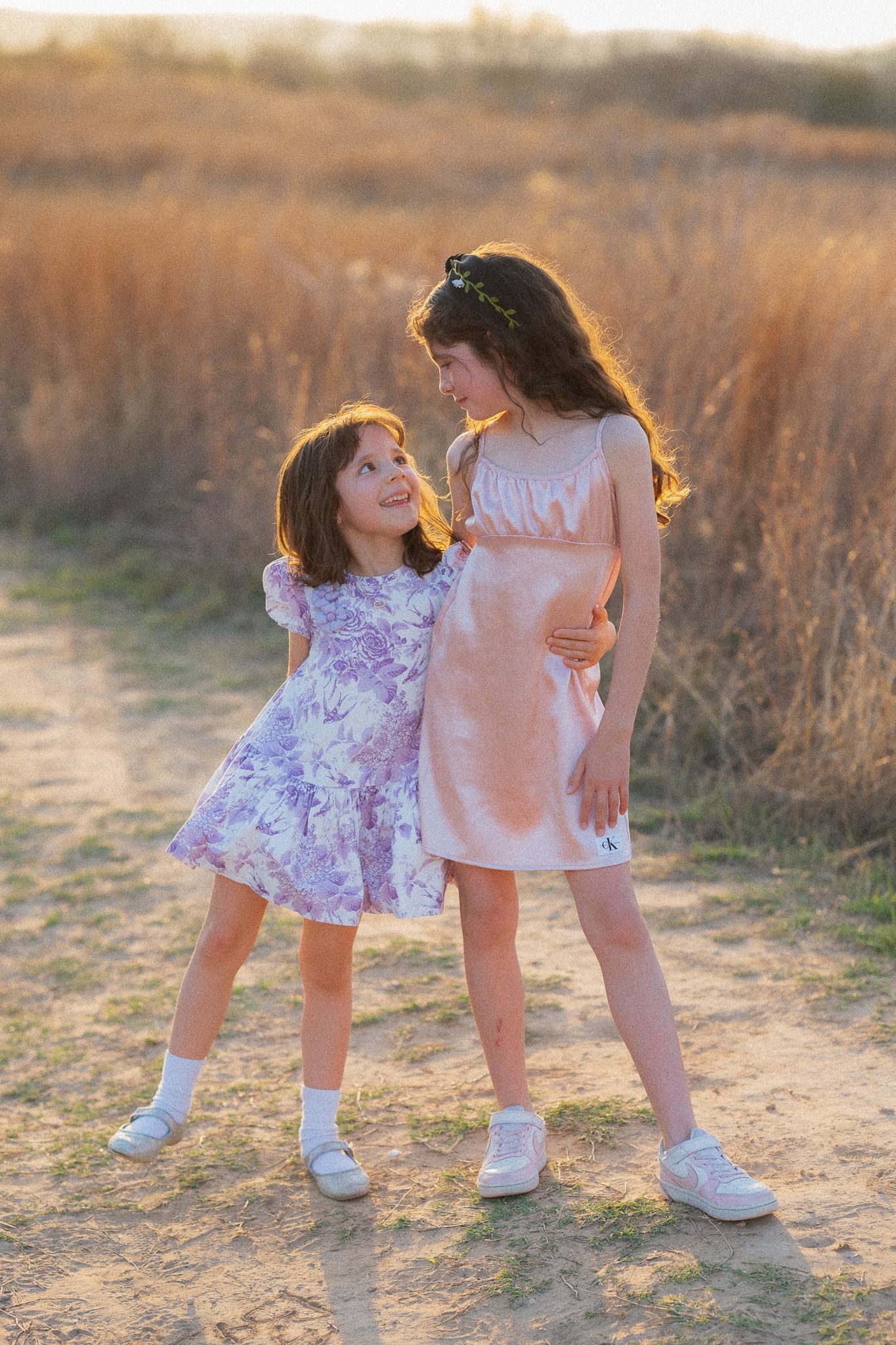 Two young girls in pastel dresses share a sweet moment on a sunny field path at golden hour.