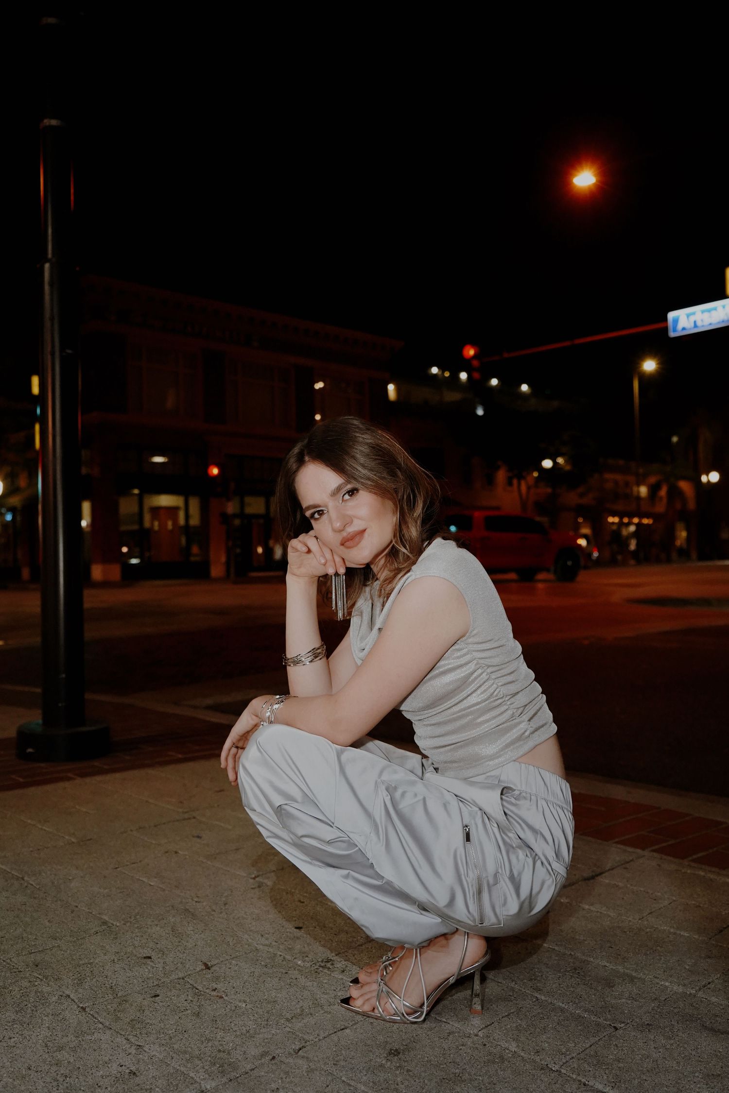 A person in white clothing crouches on a sidewalk at night with city streets and warm lighting in the background.