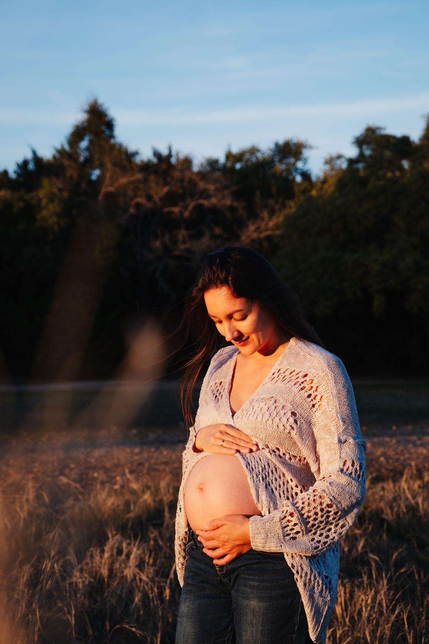 A maternity photo showing an expectant mother in a white lace sweater standing in a sunlit field.