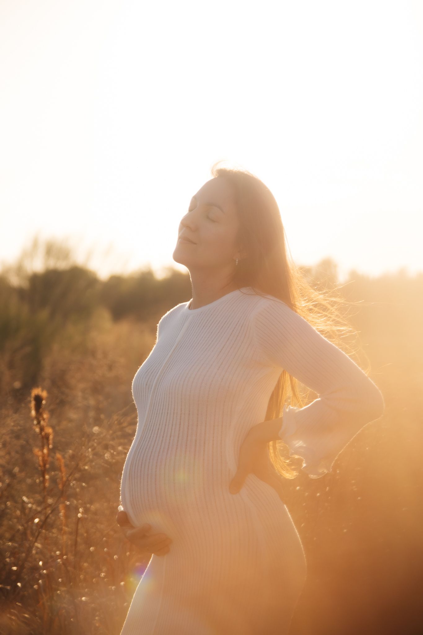 A pregnant figure in white dress poses against warm sunset lighting in a natural outdoor setting.