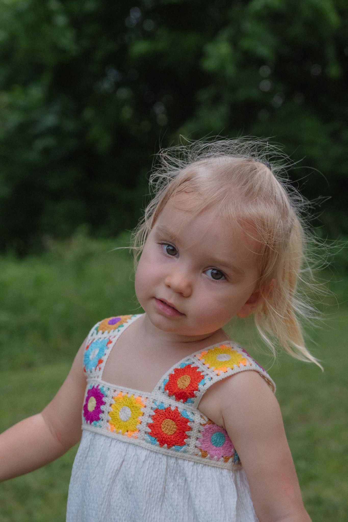 A sweet summer dress with colorful floral crochet details adorns a young subject against a natural green backdrop.