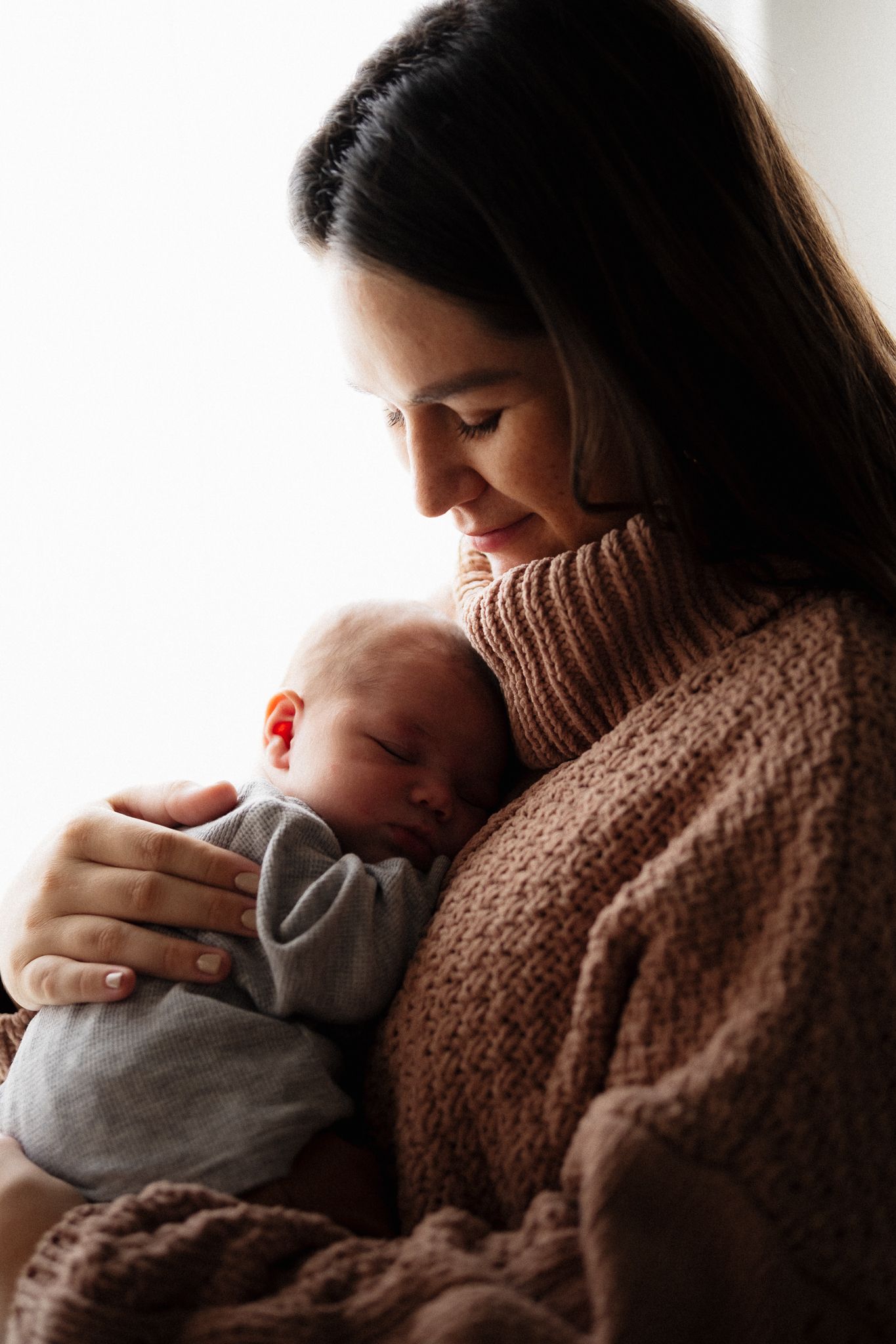 A smiling mother in a brown knit sweater tenderly holds her sleeping newborn baby close to her chest.