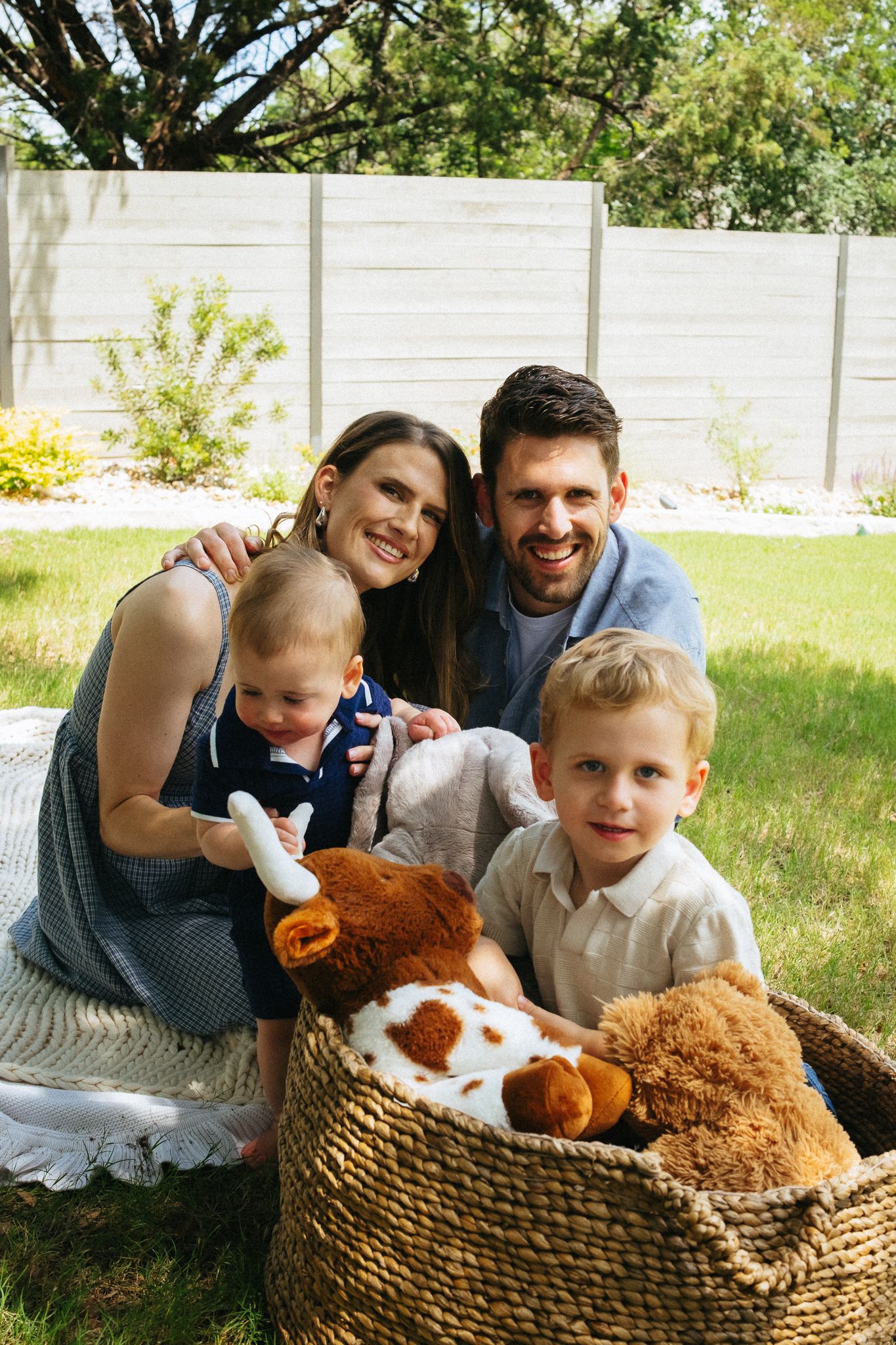 Happy family of four posing in backyard with basket of stuffed animals on sunny day.