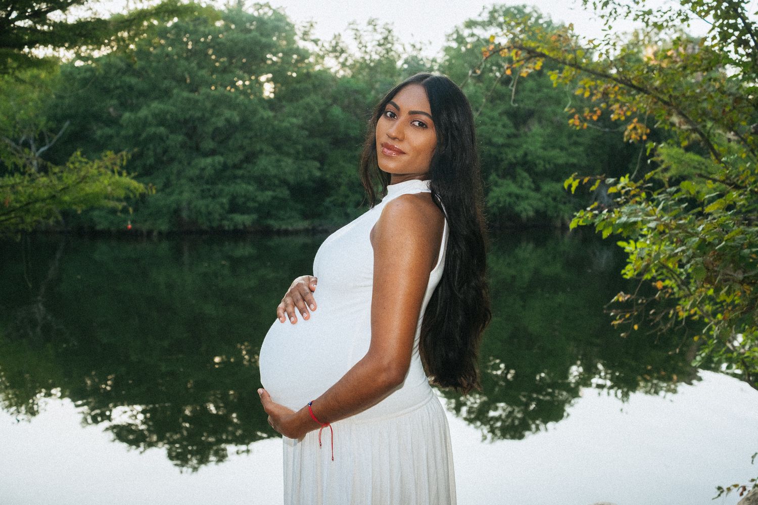 Pregnant woman in white dress holding belly by reflective lake with lush green trees backdrop.