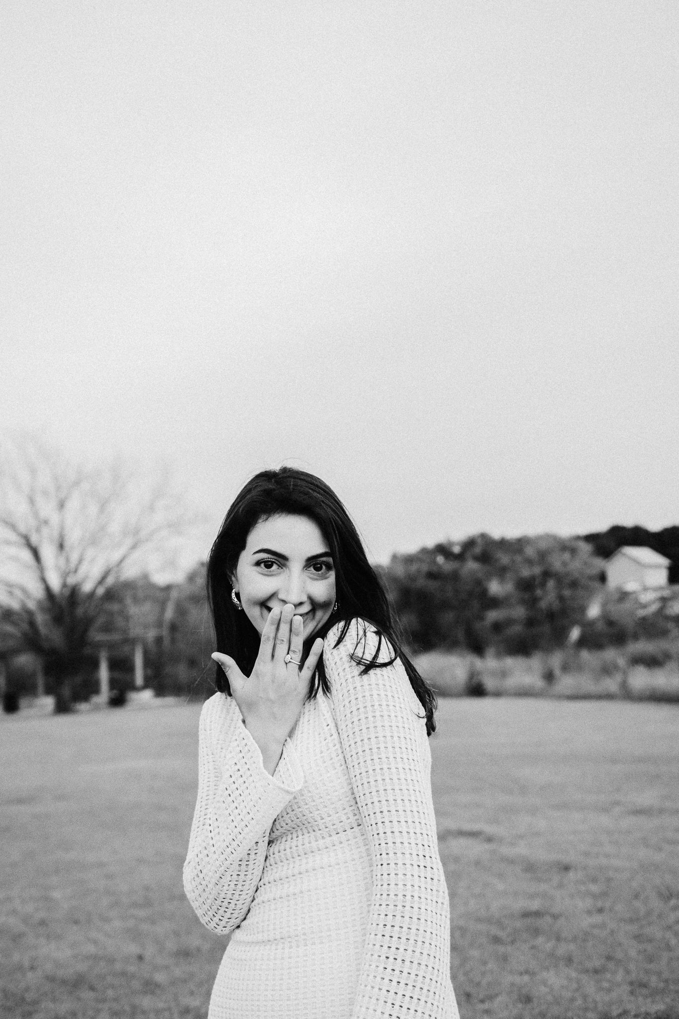 A person in a white sweater poses playfully with hand over mouth in a black and white outdoor portrait.