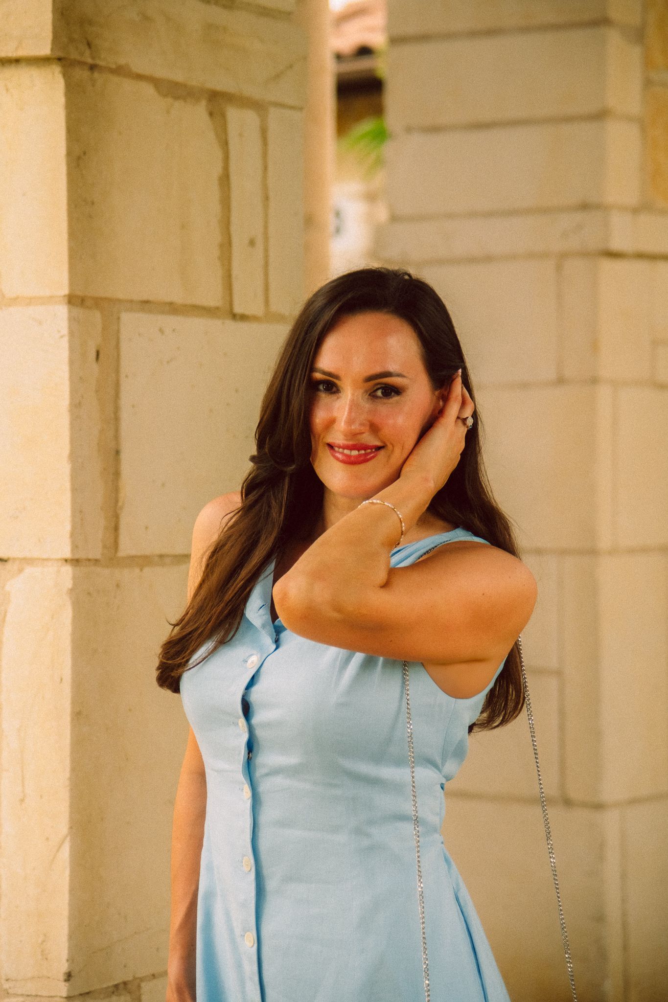 A person in a light blue dress smiles and poses against a white stone wall with columns.