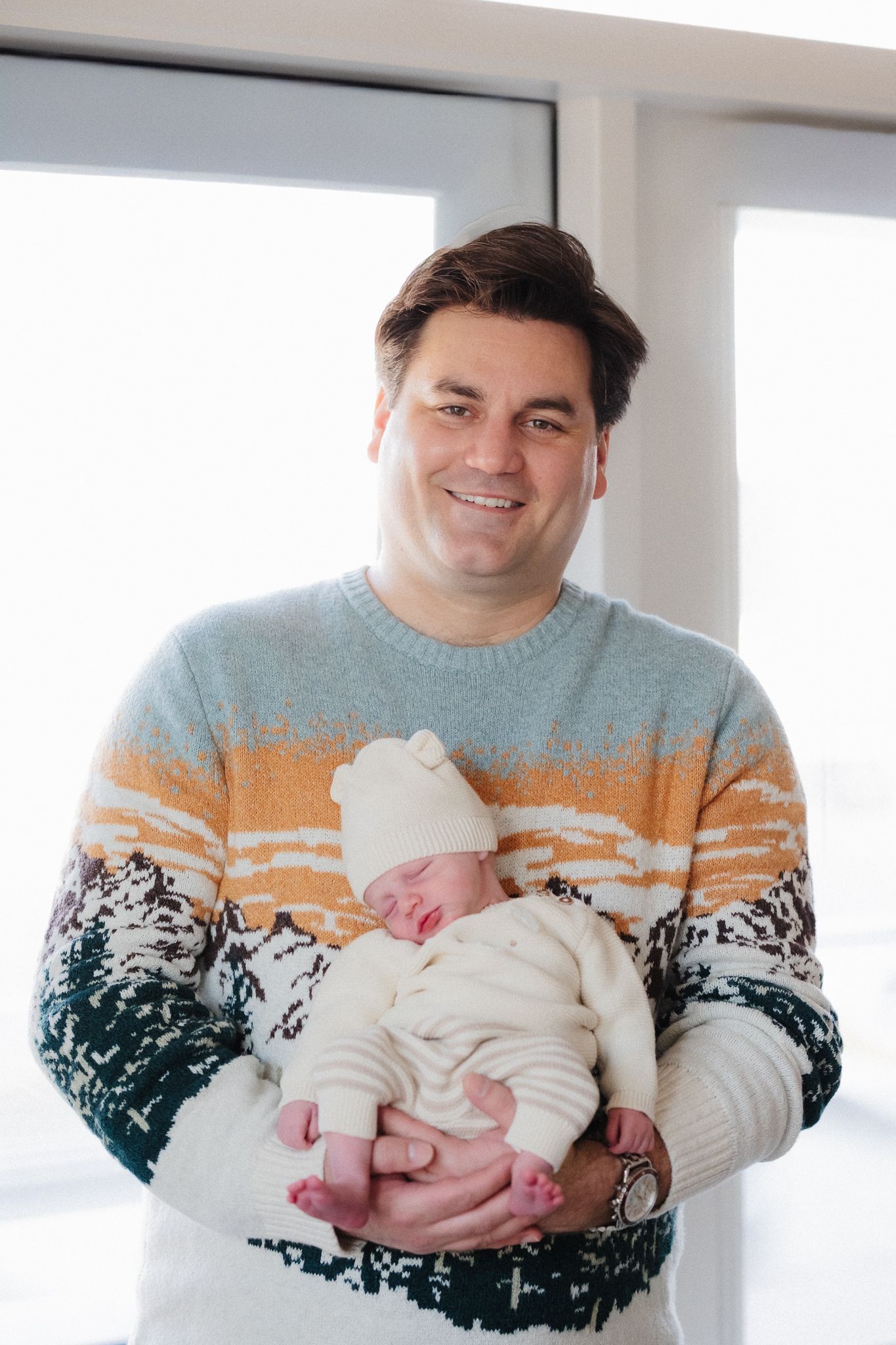 A person wearing a winter-themed sweater holds a sleeping newborn baby wrapped in white clothing.
