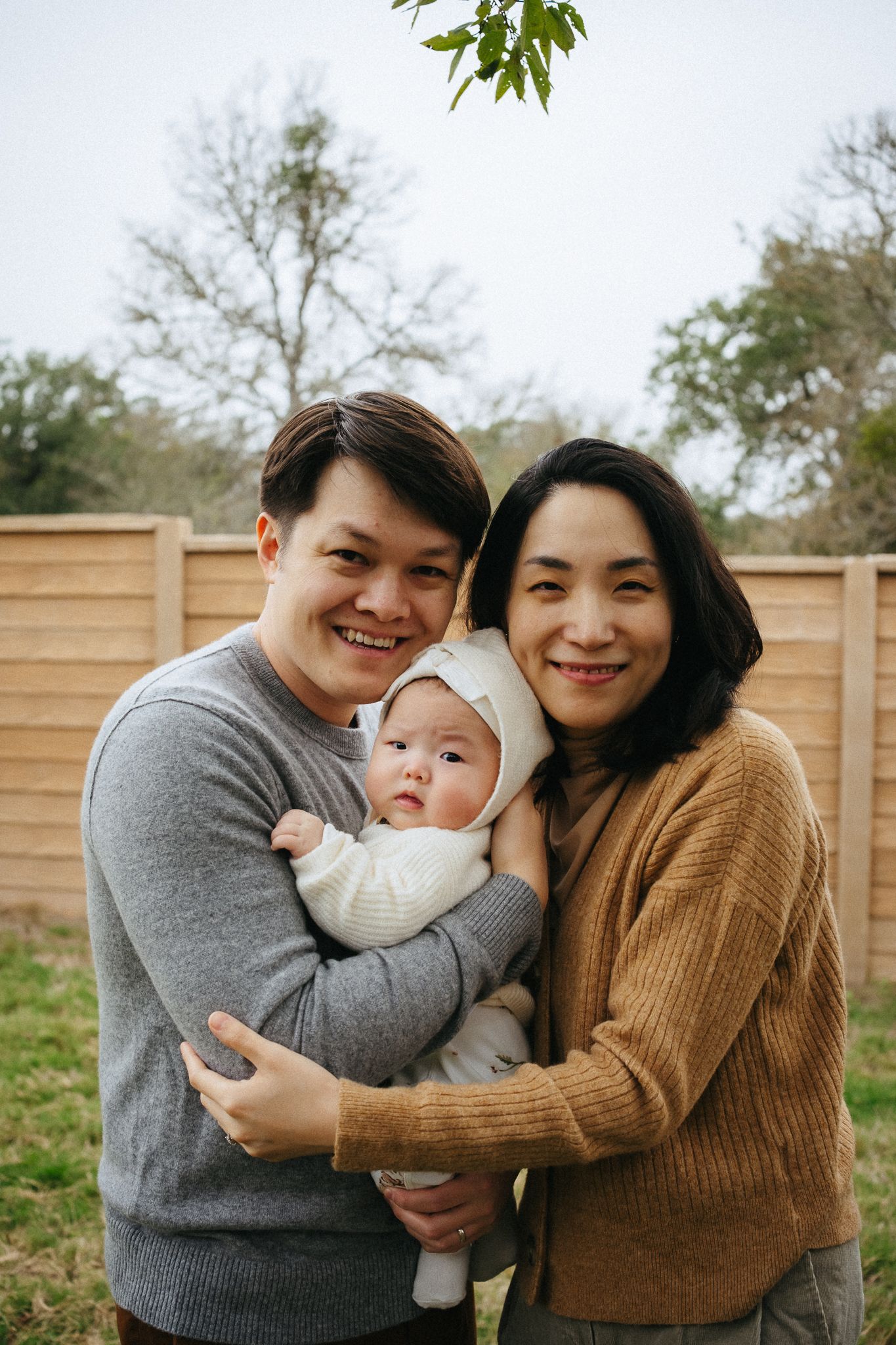 A family poses for intimate portraits outdoors against a wooden fence backdrop.