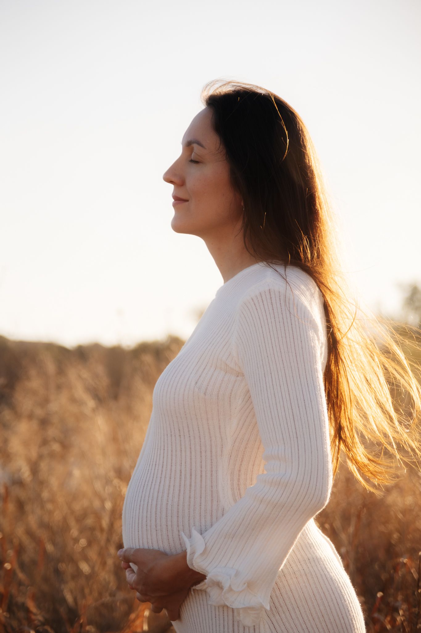 A profile view of someone in a white sweater standing in a sunlit field at golden hour.