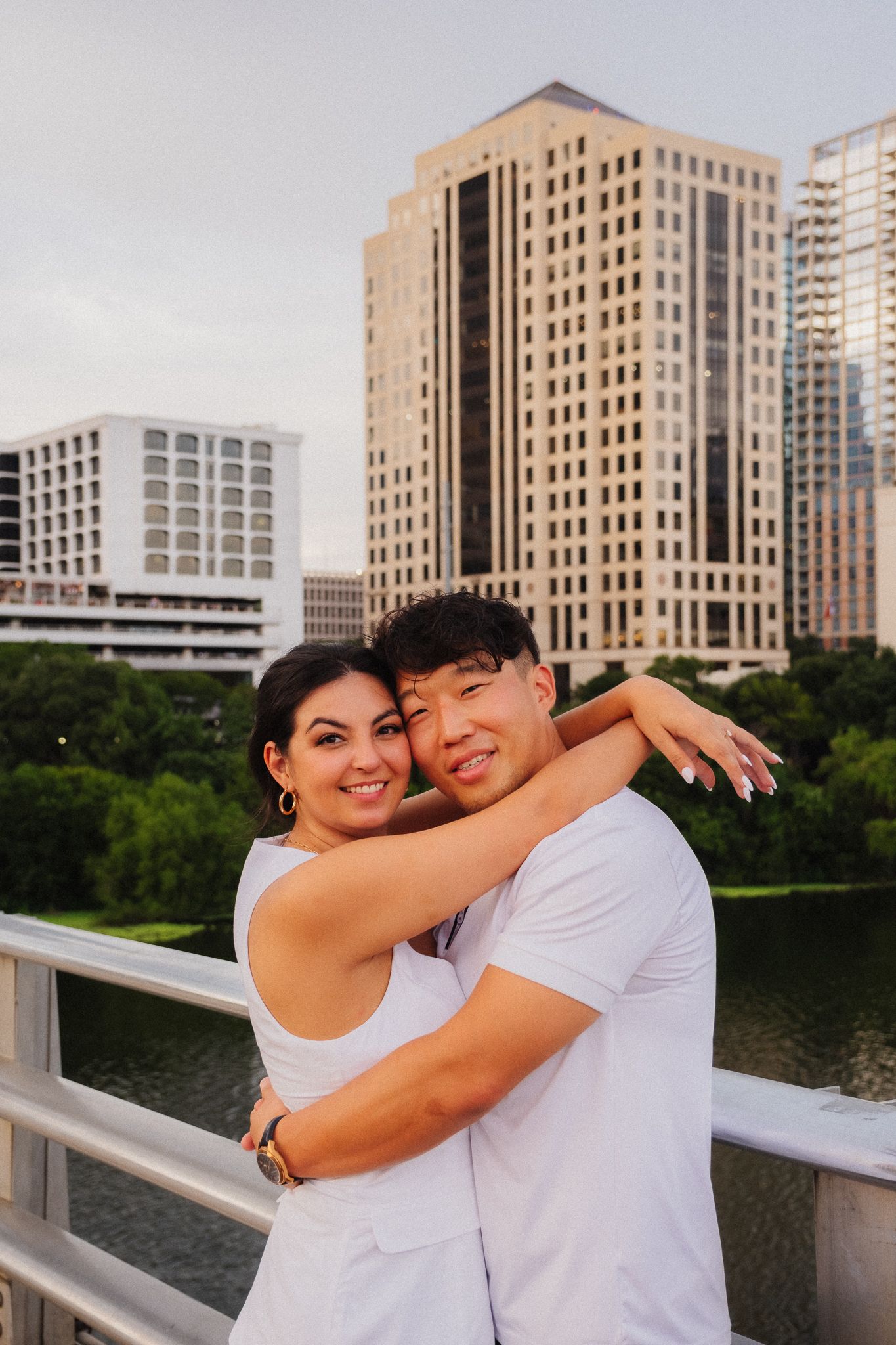 Couple in white outfits embracing on a bridge with a city skyline and green trees in the background.