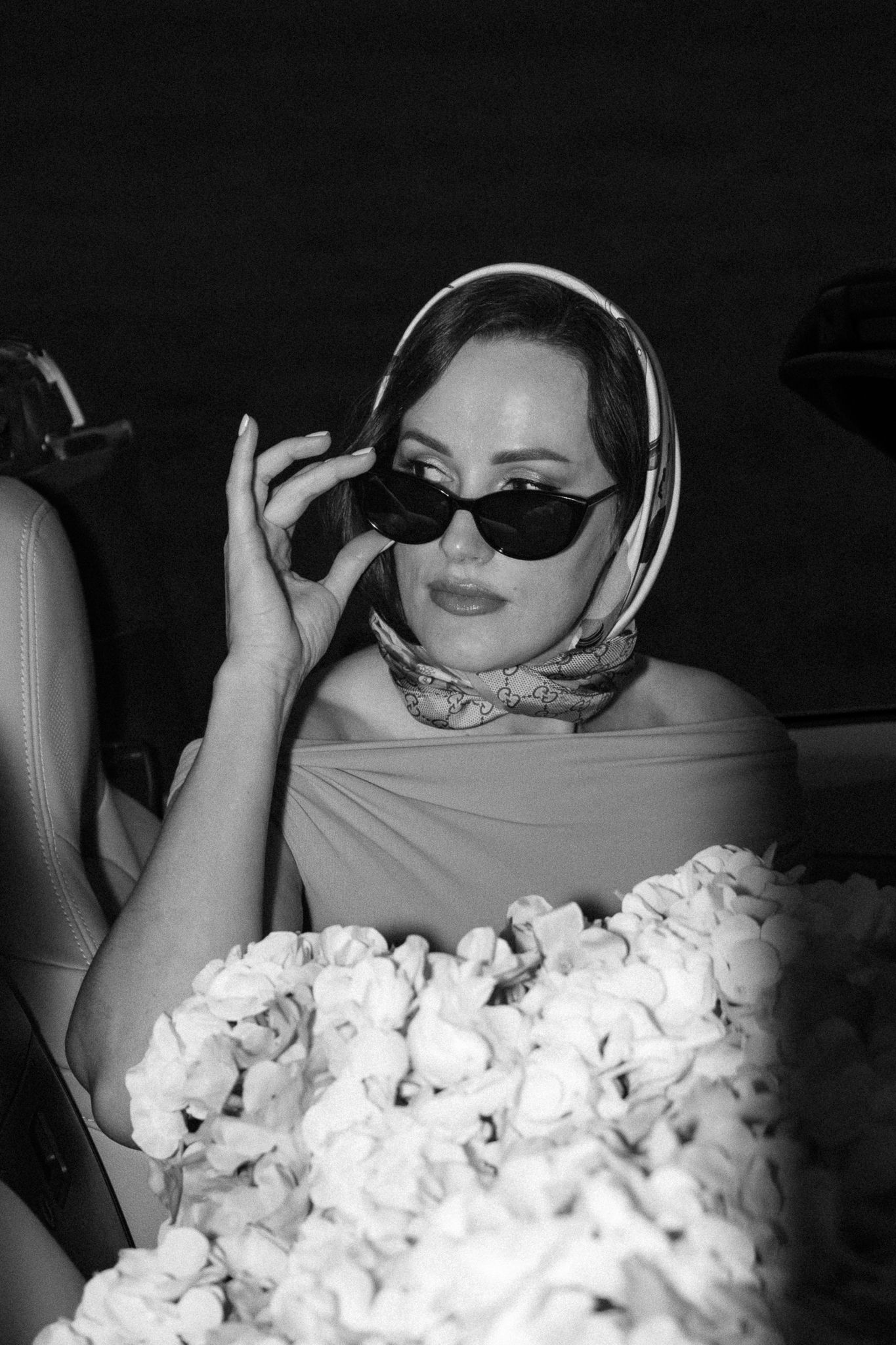 Black and white photo with someone posing in car surrounded by white flowers.