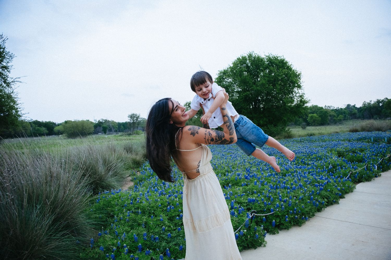 Mother in white dress lifting laughing toddler above bluebonnet field in Texas spring outdoor photo.