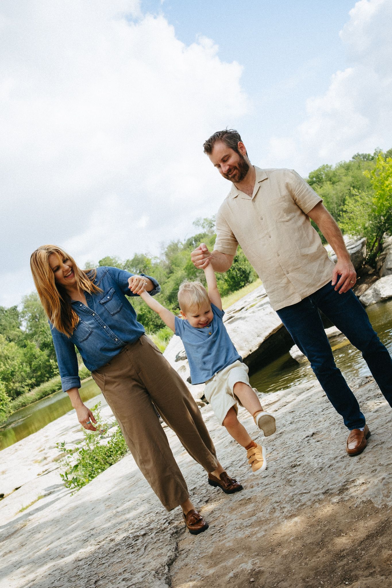 A joyful family swings their young child between them while walking on rocky terrain near a river.
