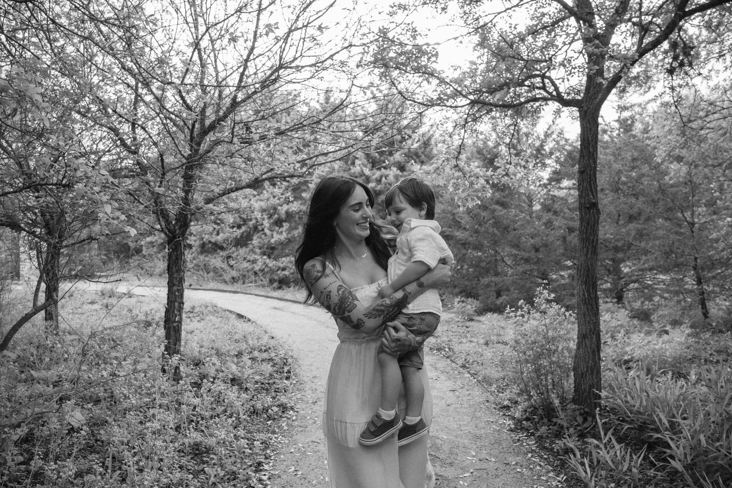 Black and white photo of a woman in a white dress holding a toddler on a tree-lined path.