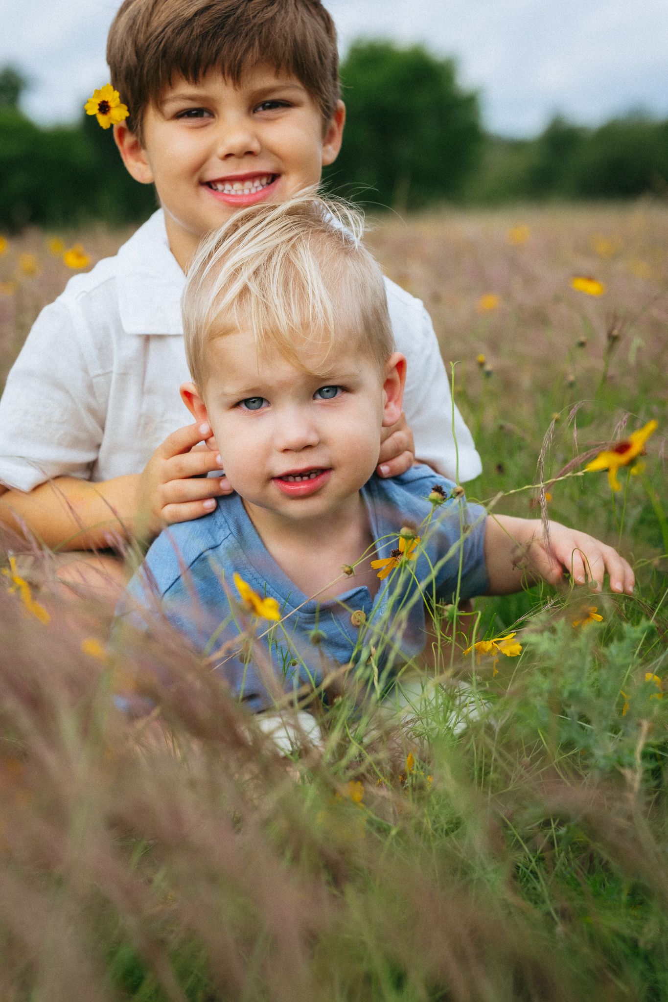 Two young boys sitting in a wildflower field, older boy smiling with a yellow flower in his hair.