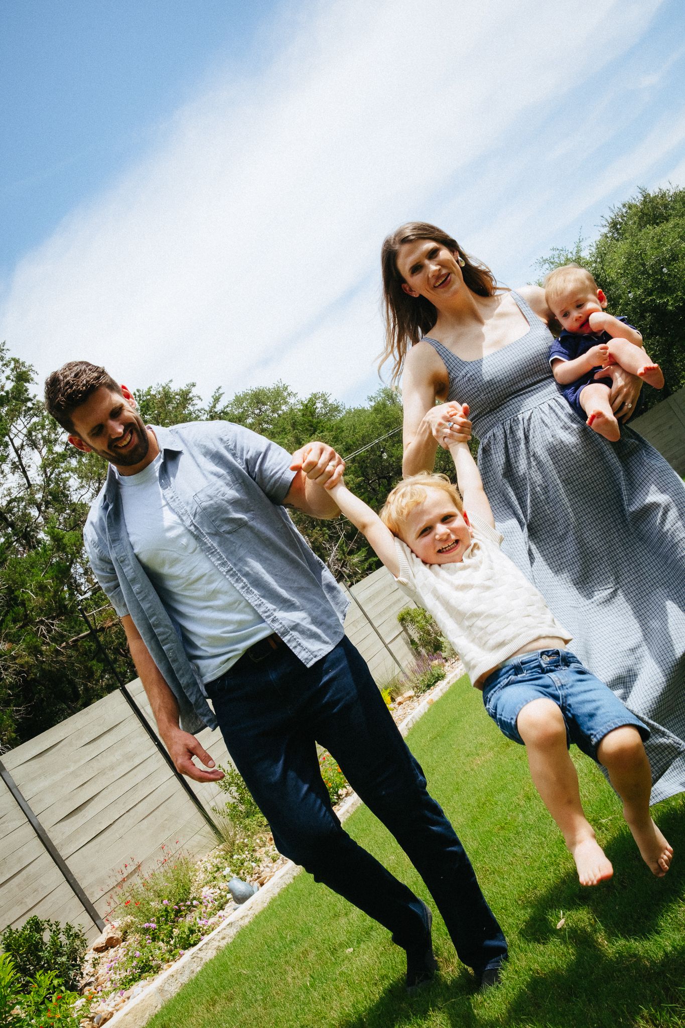 Joyful family swinging young boy in sunny backyard while mother holds baby.