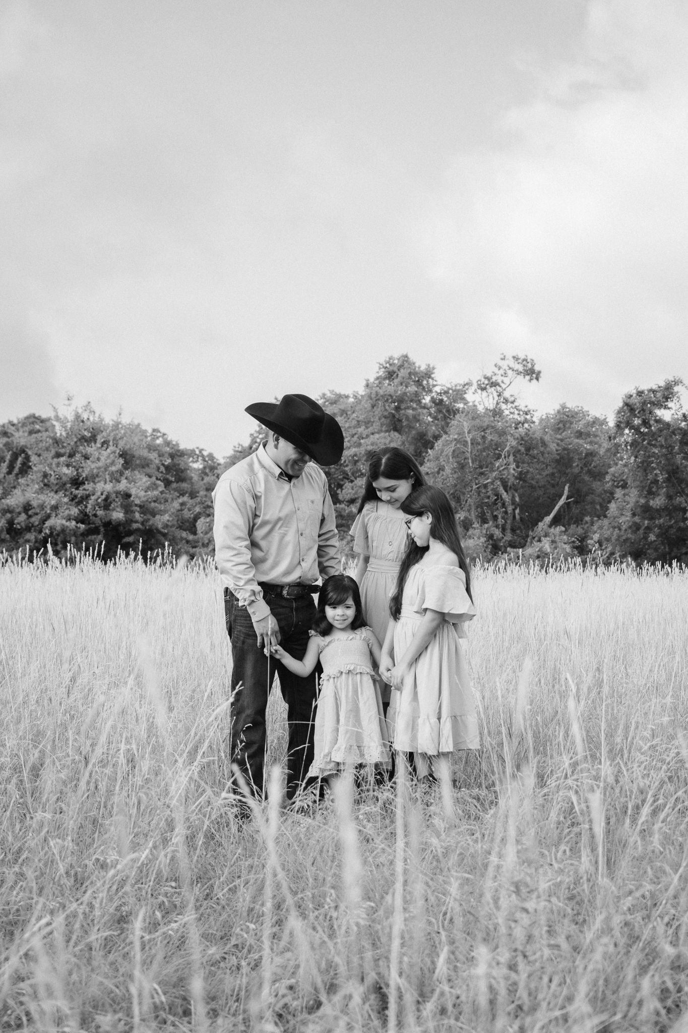 Black and white family portrait of a man in cowboy hat with three girls standing in a tall grass field.