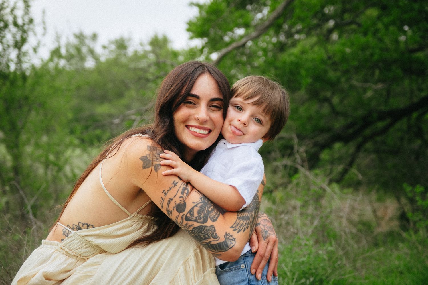 An adult and young child share a tender embrace while smiling outdoors in a natural green setting.
