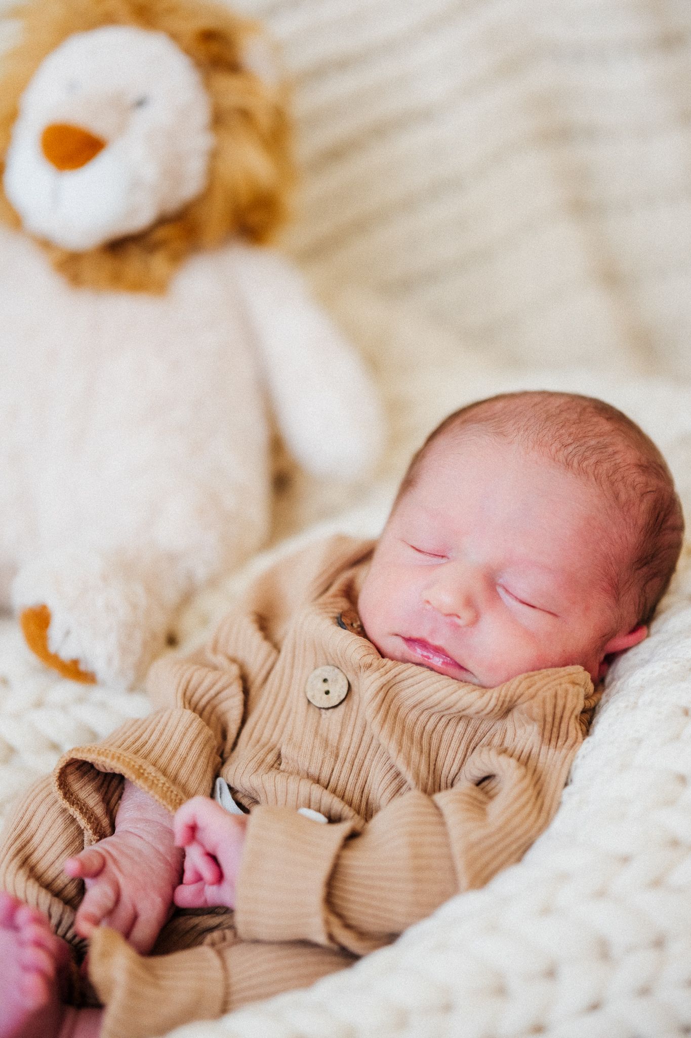 Sleeping newborn baby wearing tan knit sweater rests peacefully on white textured blanket with stuffed animals nearby.