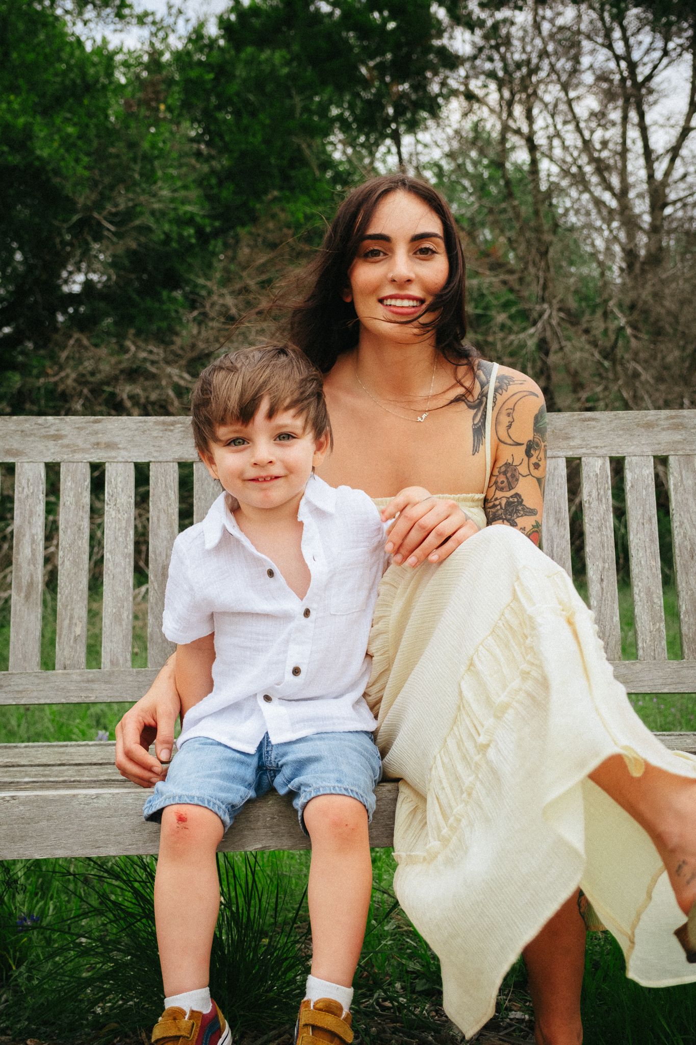 A figure in a white dress and tattoo sits on a wooden park bench next to a young child in a white shirt and shorts.