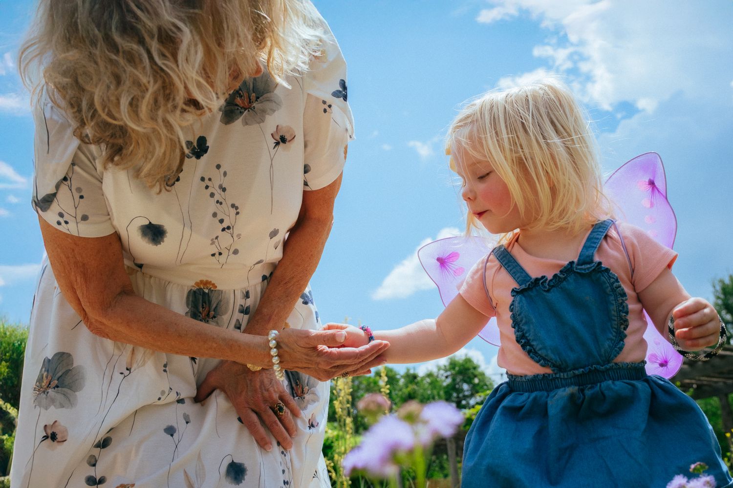 Young girl in fairy wings receives something from a woman in a floral dress in a sunny garden.