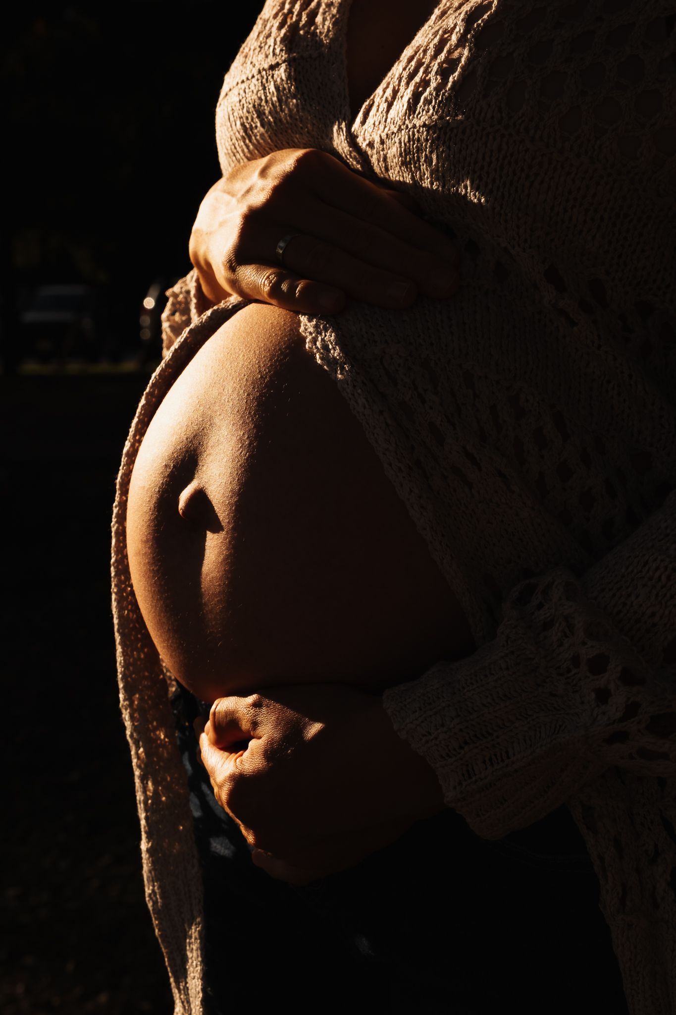 Side view of a pregnant belly silhouetted in dramatic lighting with hands gently resting on the baby bump.