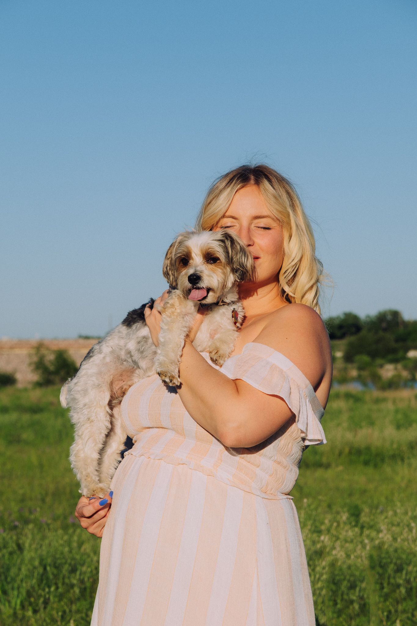 A small fluffy dog is held lovingly in a grassy field by someone wearing a flowy pink off-shoulder dress.