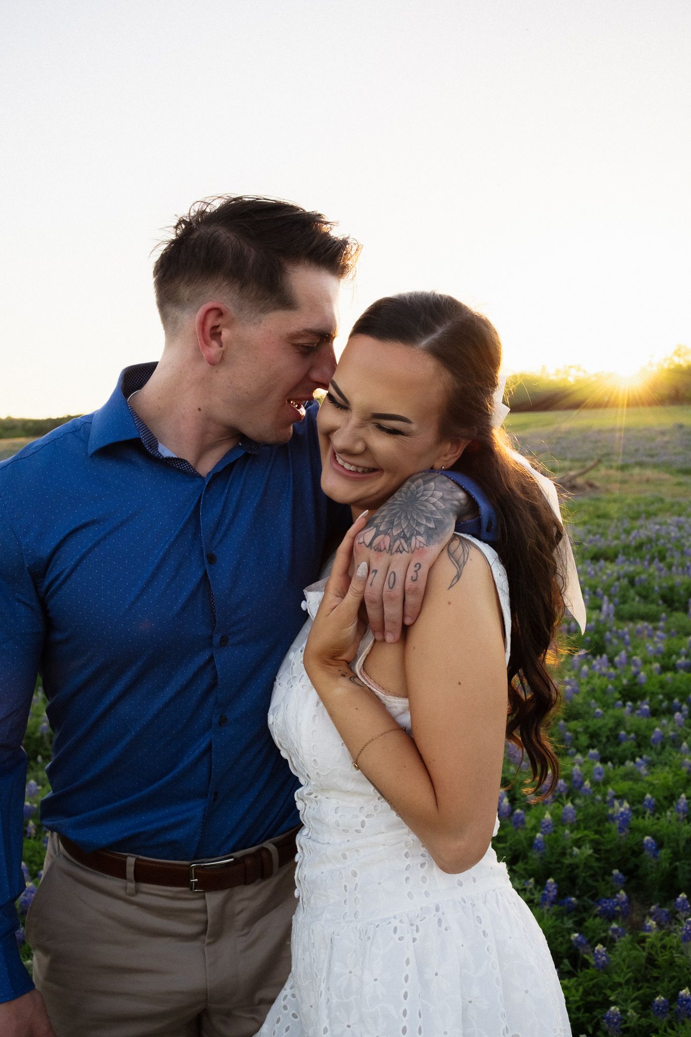 Couple embracing in a field of bluebonnets at golden hour, woman in white dress laughing joyfully.