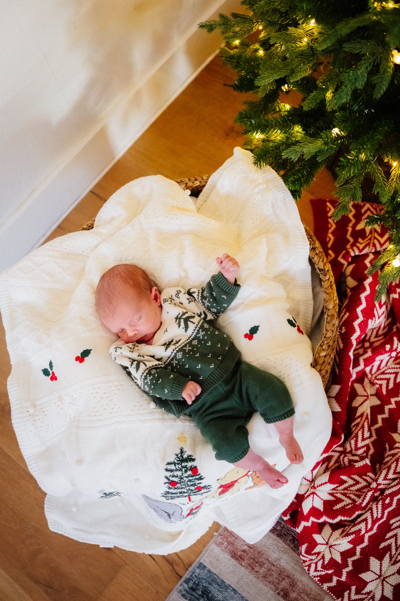 Newborn baby sleeping on white blanket in green Christmas outfit beside decorated tree with lights.