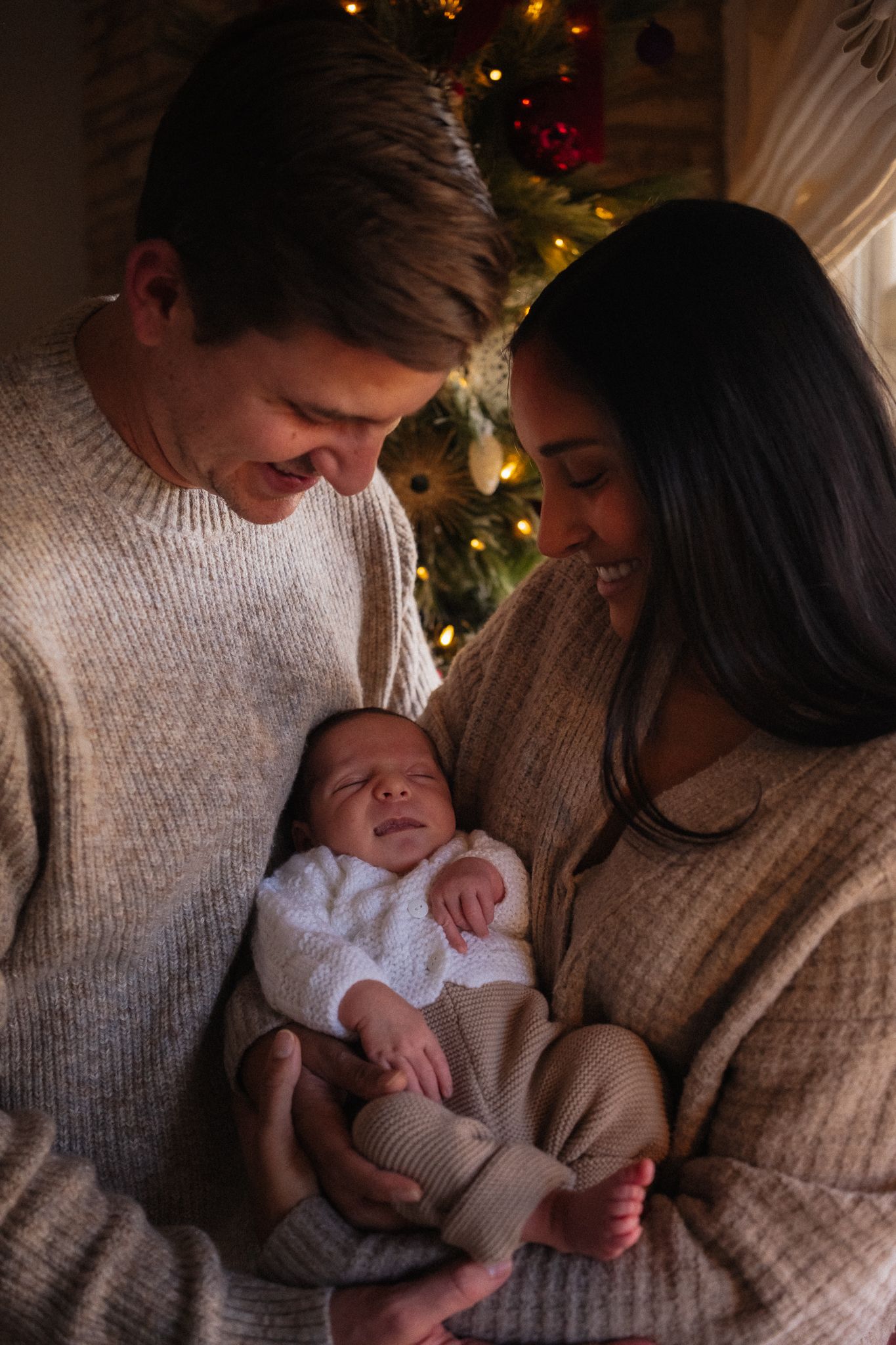 New parents holding their newborn baby in front of a Christmas tree, wearing matching beige sweaters.