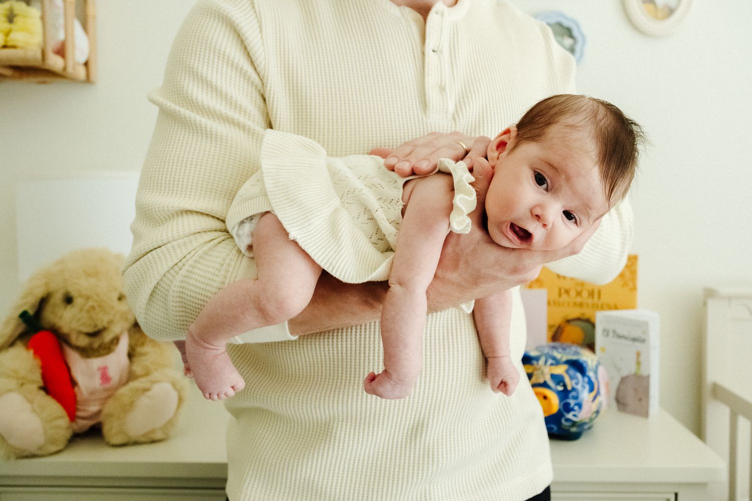 Adult holding a smiling newborn baby up in a nursery room with stuffed animals and books in background.
