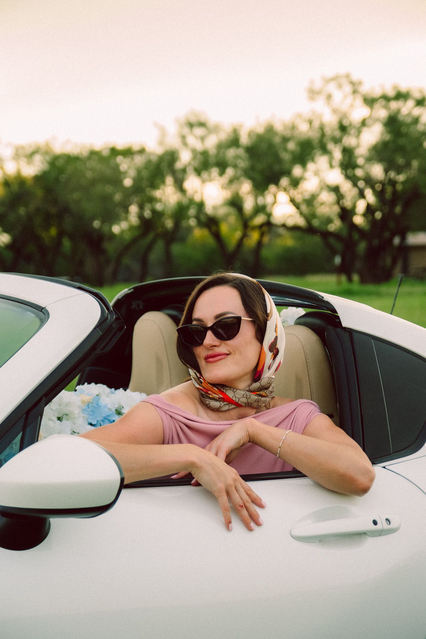 A person wearing sunglasses and a headscarf sits in a white convertible car on a sunny day with trees in the background.