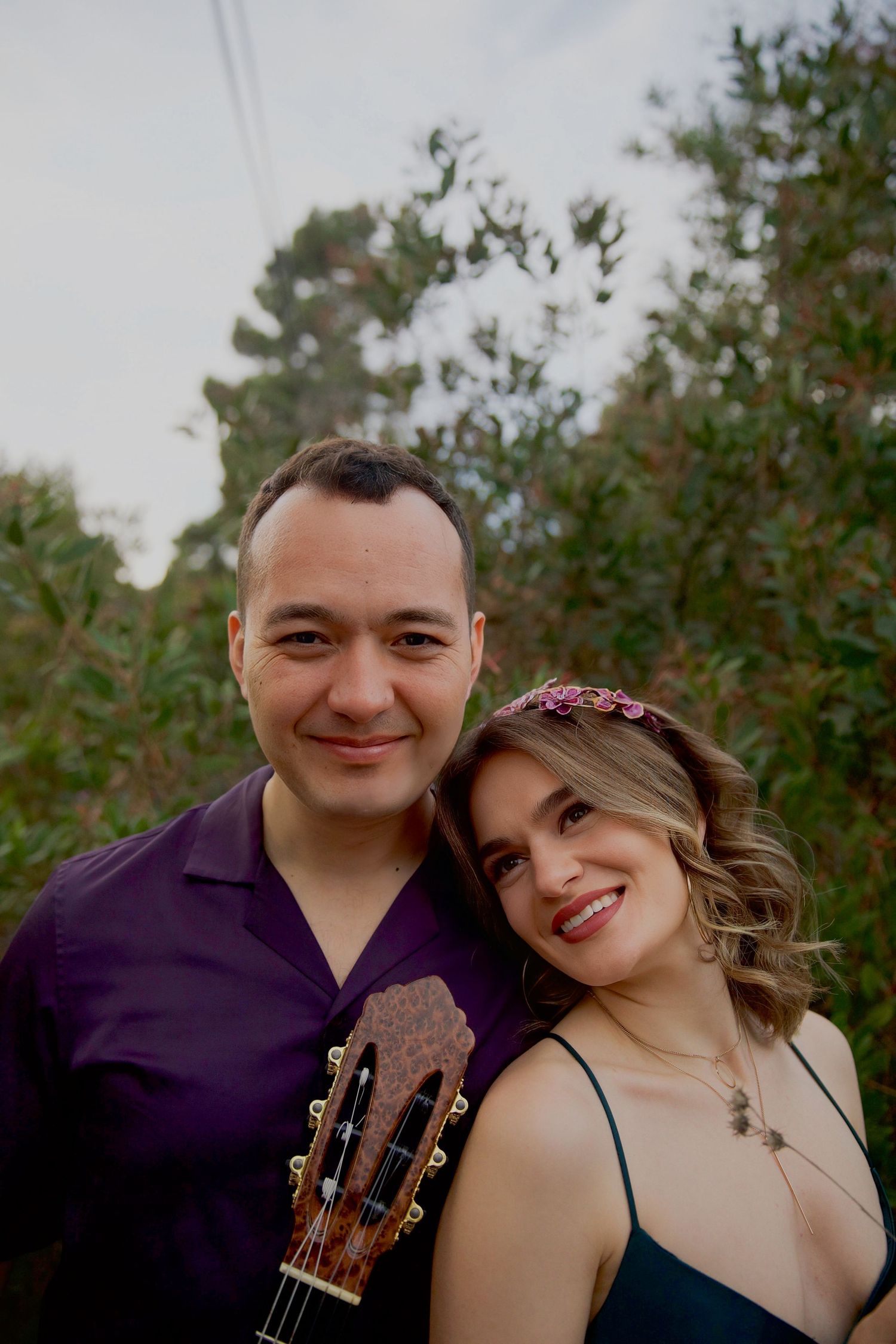 Two musicians pose outdoors with a classical guitar against a natural background with trees and greenery.