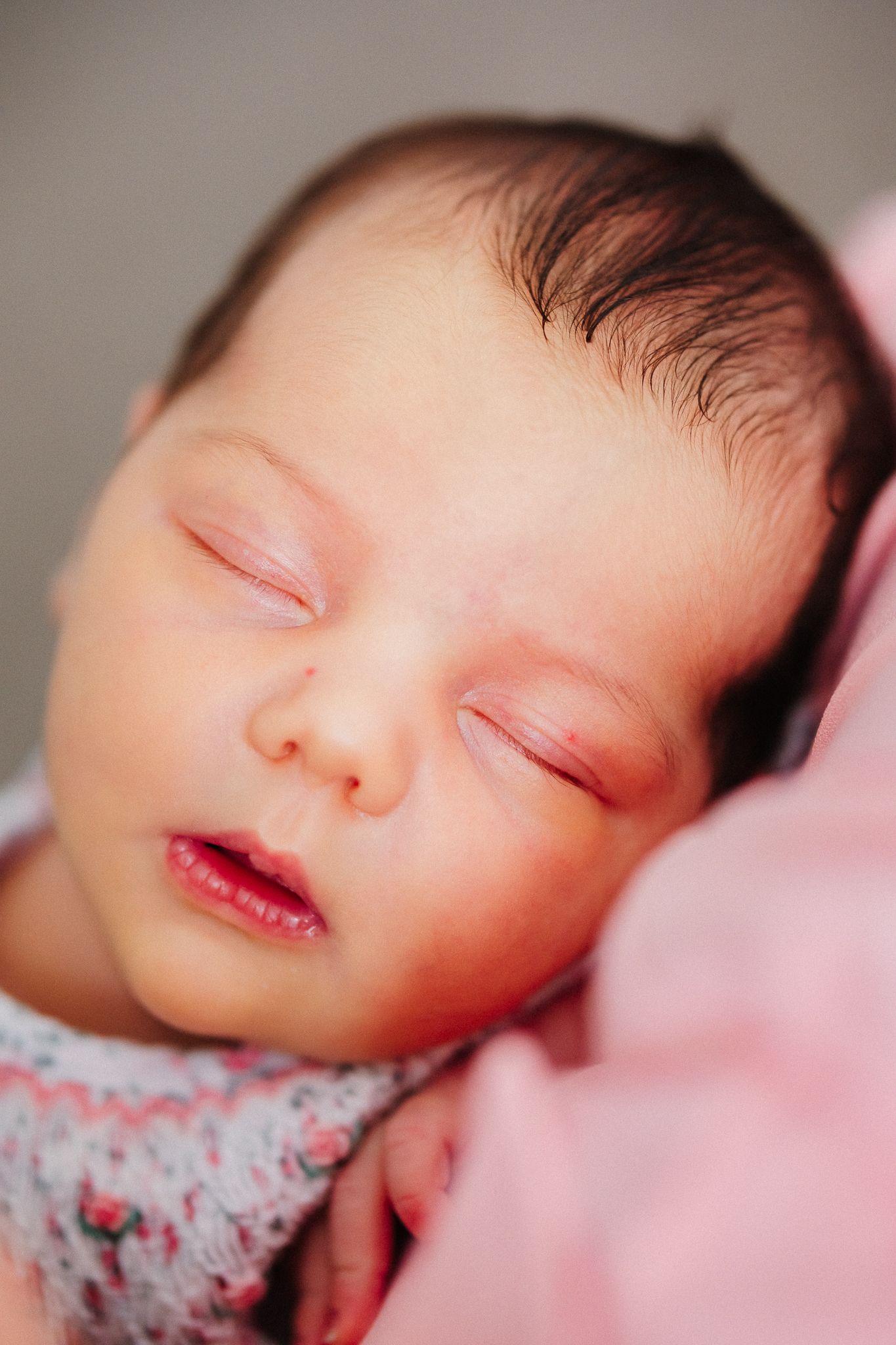 Peaceful sleeping newborn wearing floral outfit rests on pink blanket in soft natural light.