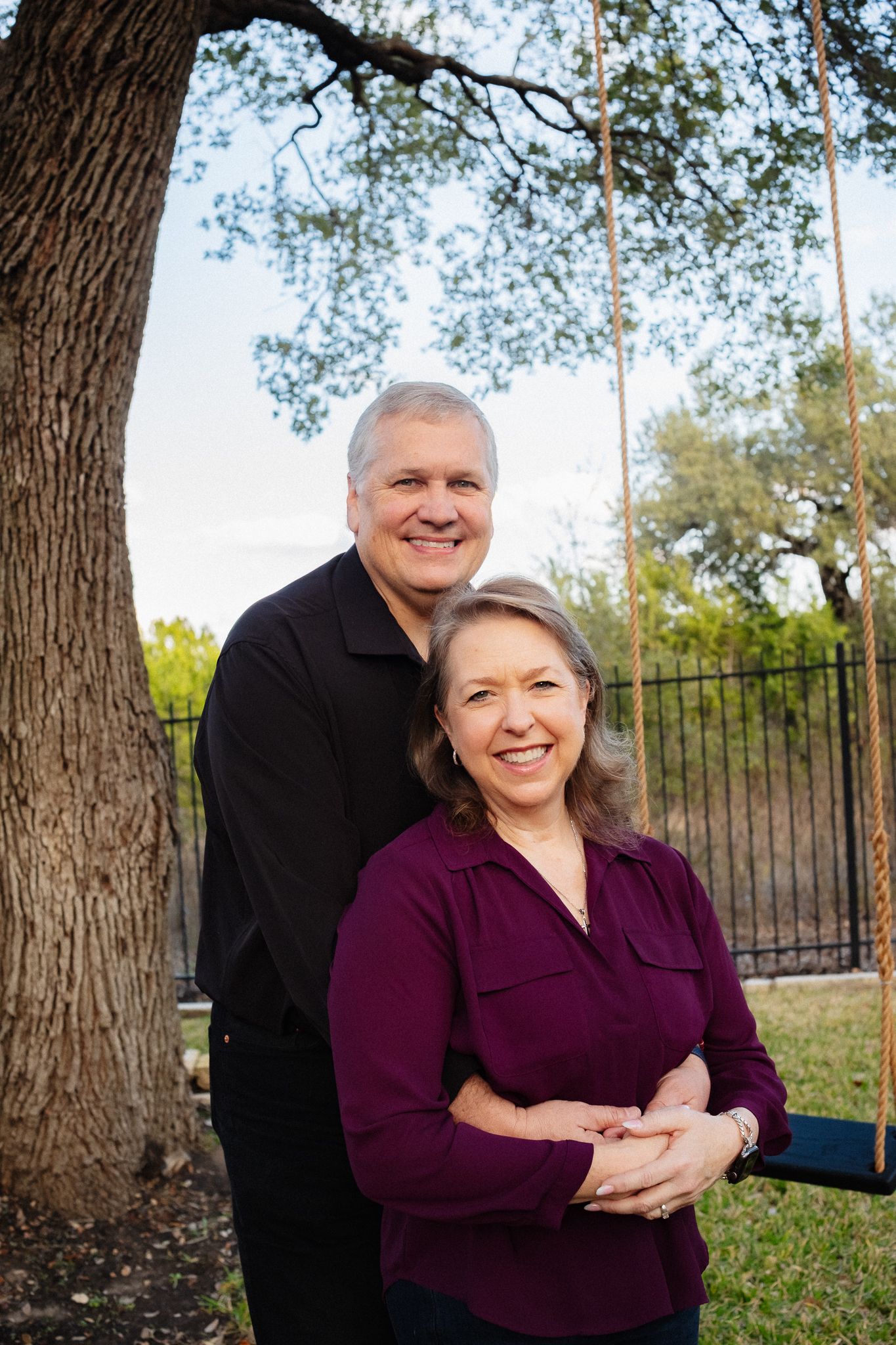 Couple poses together outdoors with trees and iron fence in background during sunset photoshoot.
