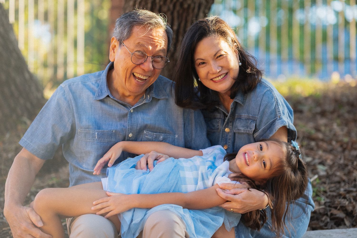 Grandparents laughing and playing with their young granddaughter outdoors on a sunny day.