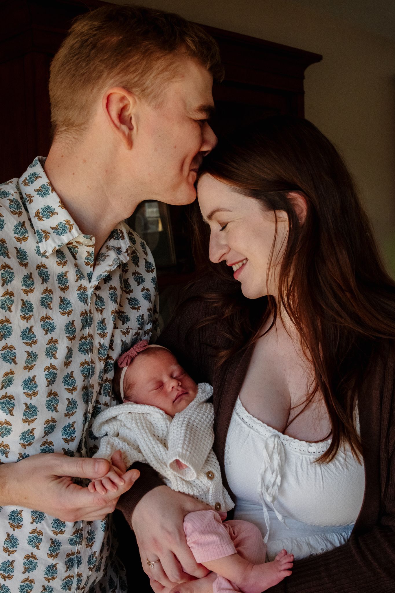 A young couple smiles at each other while lovingly holding their newborn baby dressed in white.