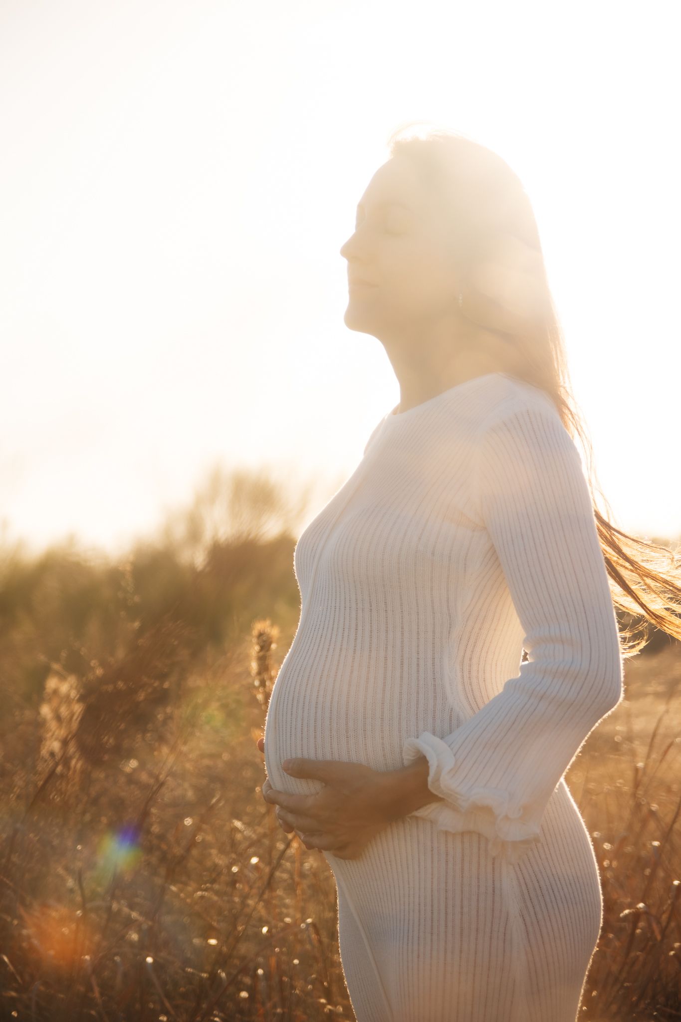 A silhouetted pregnant woman in a white sweater dress stands in a sunlit field during golden hour.