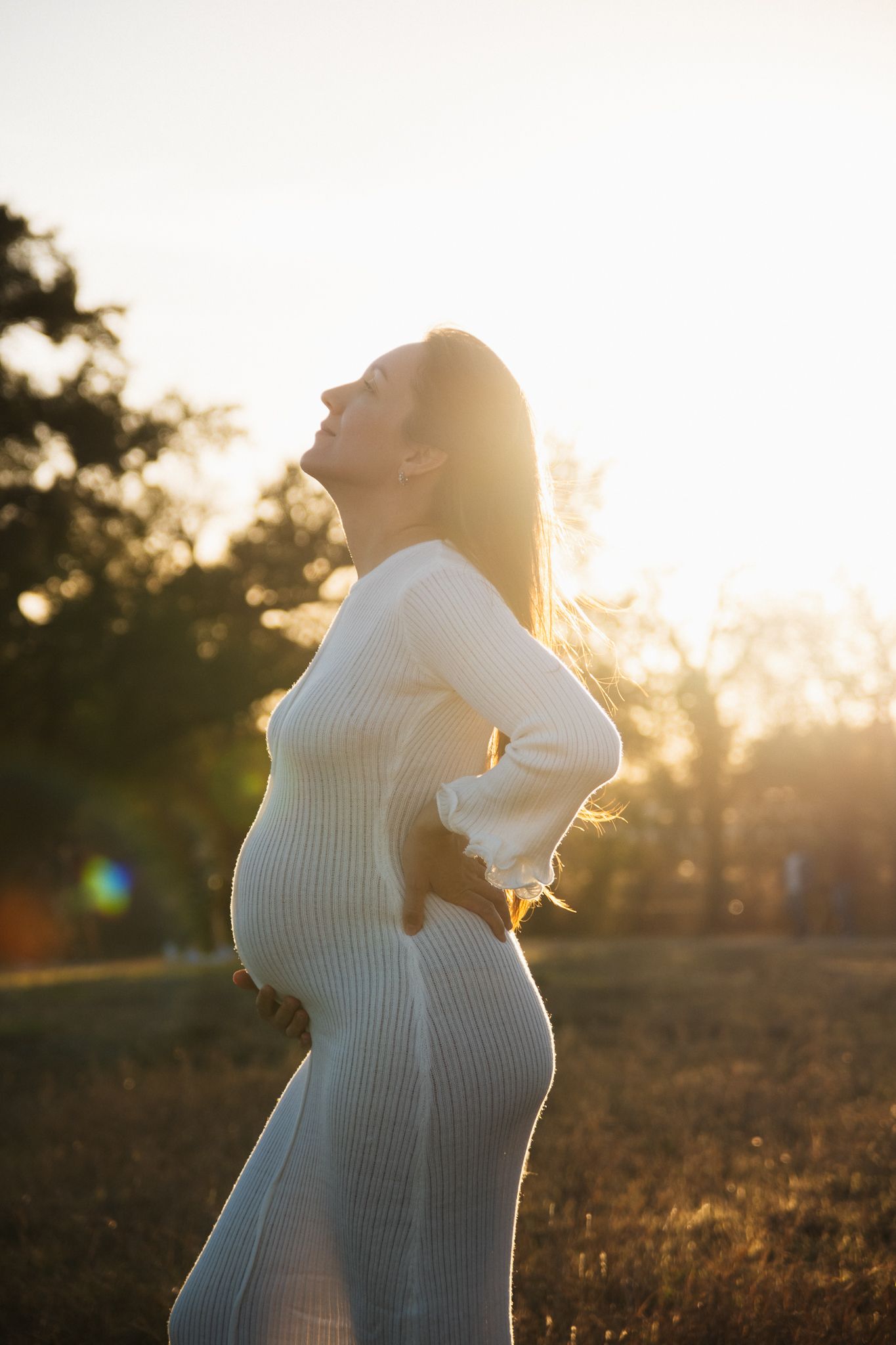 Silhouette of pregnant woman in white dress standing in golden sunlight during sunset in nature.