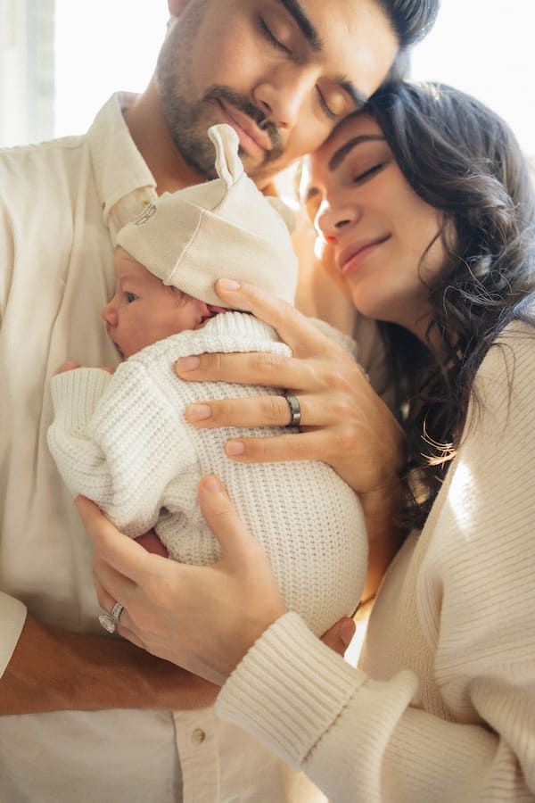 A couple tenderly holds their newborn baby wrapped in white, sharing a peaceful moment with eyes closed.